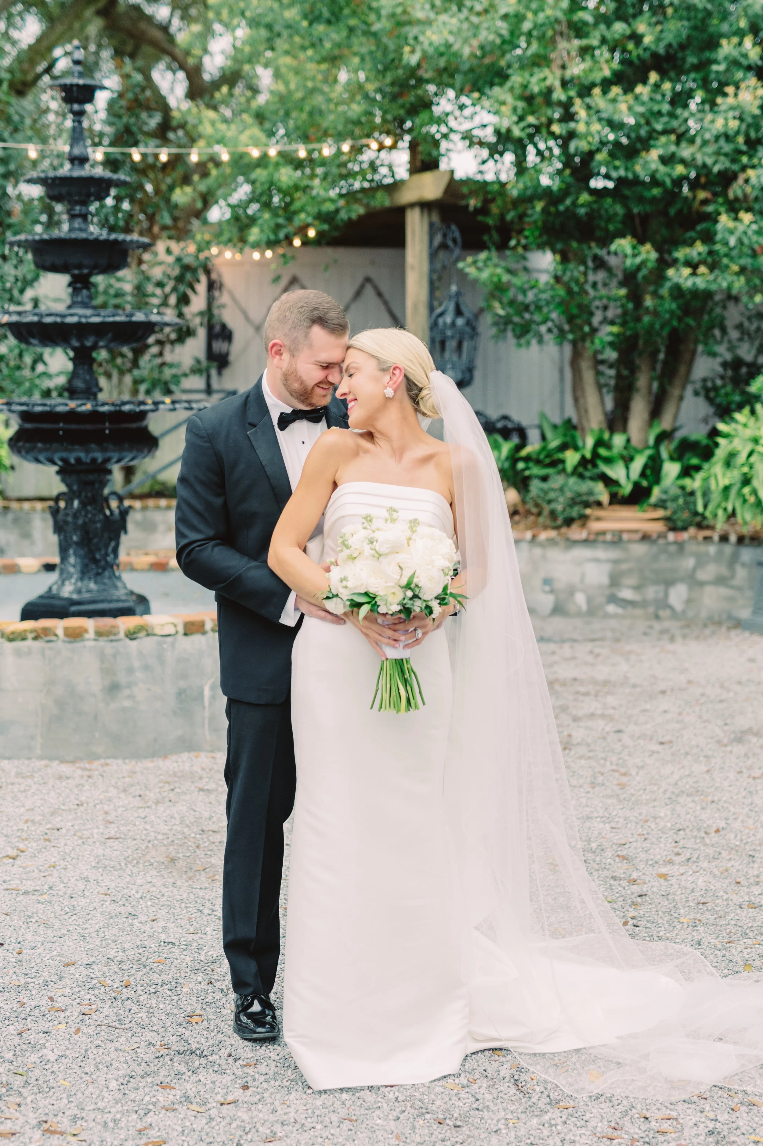 A bride and groom are smiling and leaning close together at their wedding outdoors, with trees, a fountain, string lights, and plants in the background.