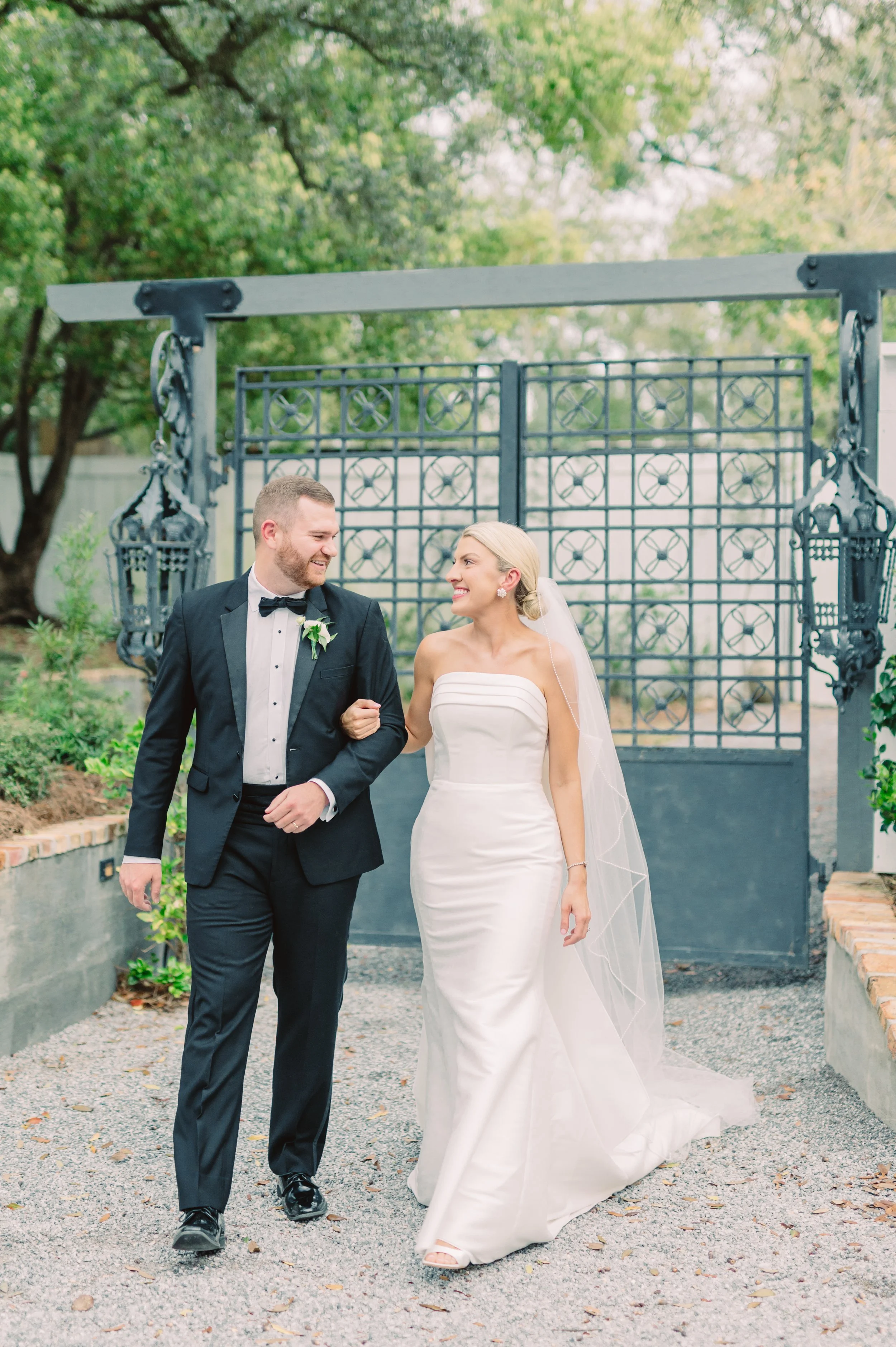 A bride and groom walking arm in arm outdoors, smiling at each other, with a decorative metal gate and trees in the background.