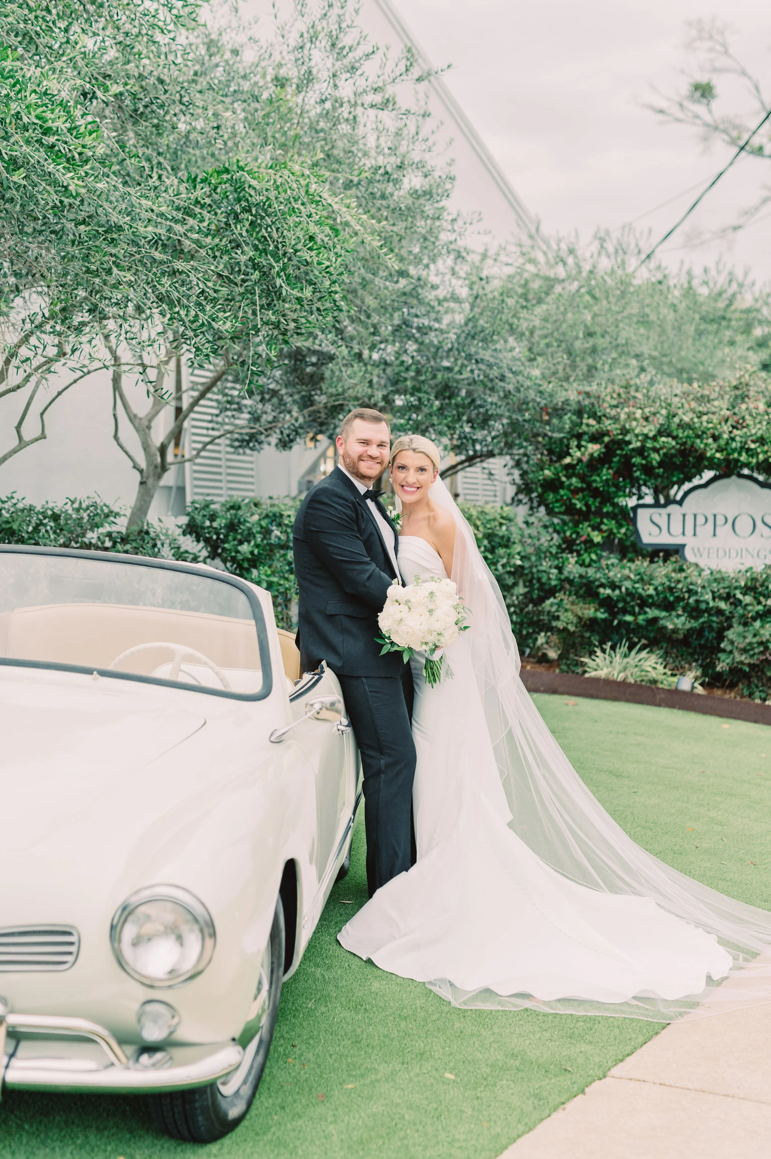 A bride and groom standing beside a vintage white convertible car outdoors during their wedding. The bride is wearing a strapless white wedding gown with a long veil, holding a bouquet of white flowers. The groom is in a black tuxedo with a bow tie. 