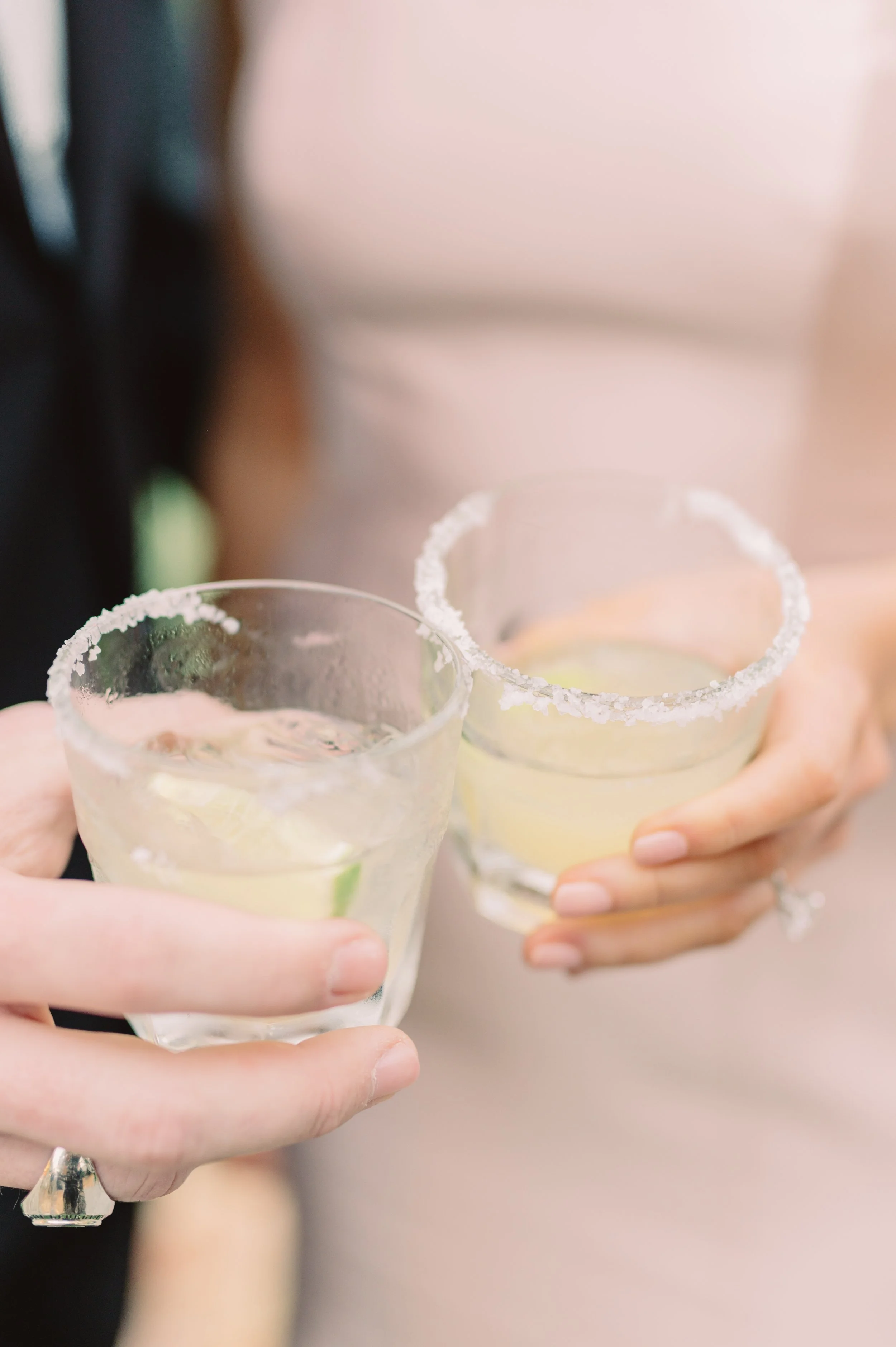 Two people holding glasses of margarita with salt-rimmed edges, lime wedges inside, and ice cubes, in a close-up shot.