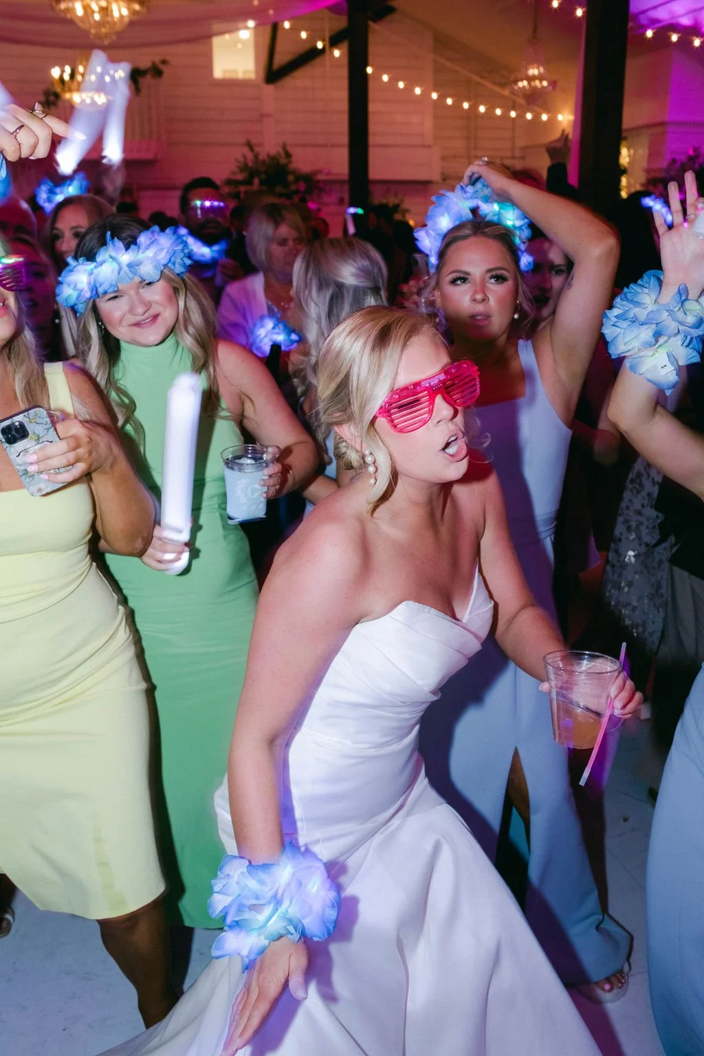 A group of people dancing at a party, many wearing floral wristbands and glow accessories, with some wearing colorful glasses and others holding drinks, under string lights and a chandelier.