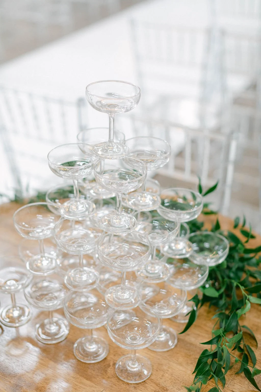 Tiered display of empty champagne coupe glasses on a wooden table with green foliage decoration in the background.