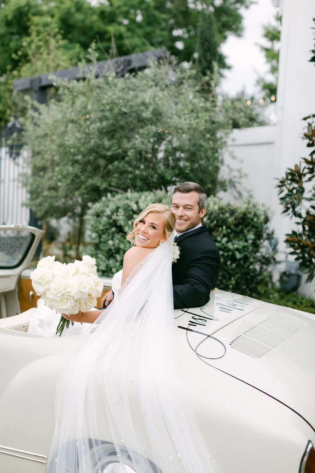 Smiling bride holding a bouquet and groom sitting in a vintage convertible car outdoors.