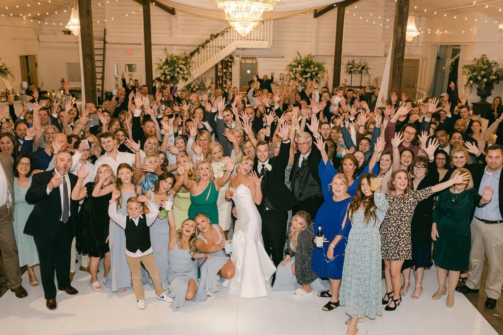 Large wedding reception with a groom and bride in the center, surrounded by guests raising their hands in celebration in a decorated venue with string lights and floral arrangements.