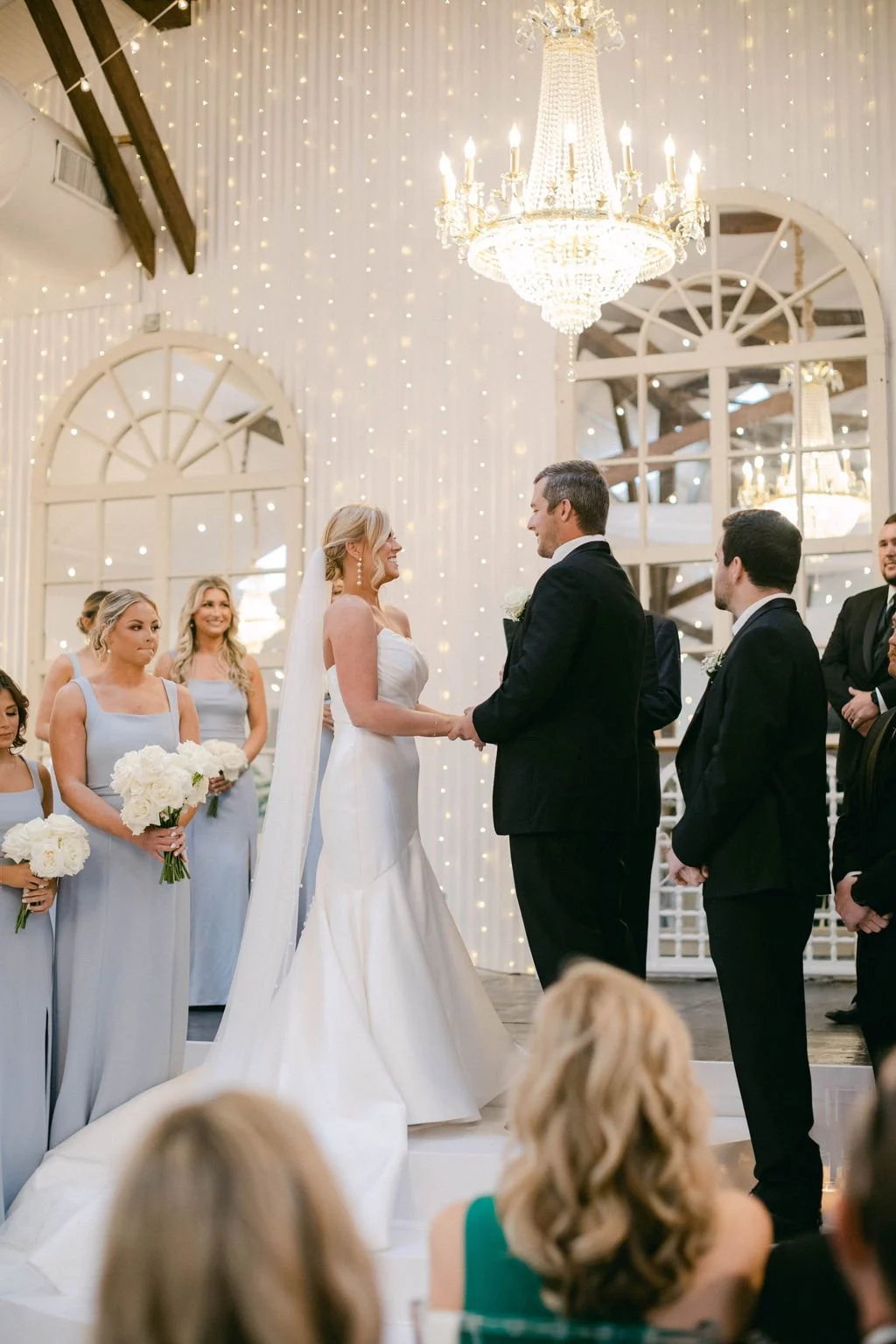 Bride and groom holding hands during their wedding ceremony in a decorated indoor venue with chandeliers and string lights, surrounded by bridesmaids and groomsmen.