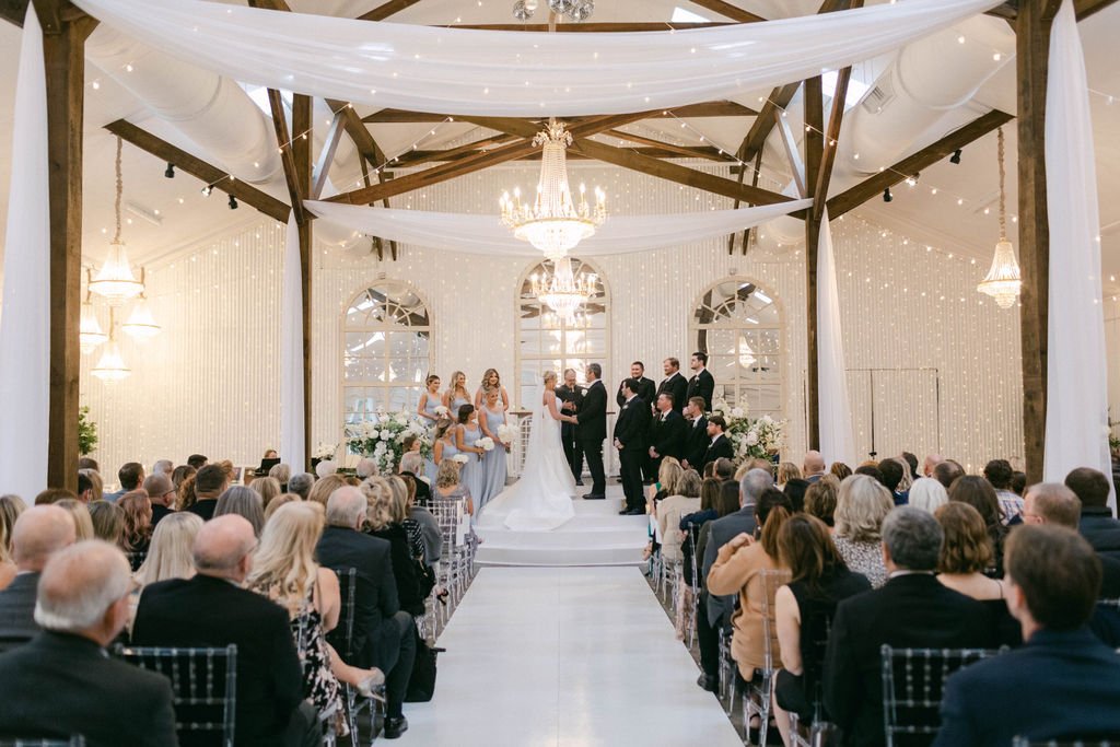 Wedding ceremony taking place inside a decorated venue with a bride, groom, bridesmaids, and groomsmen, and guests seated in rows.