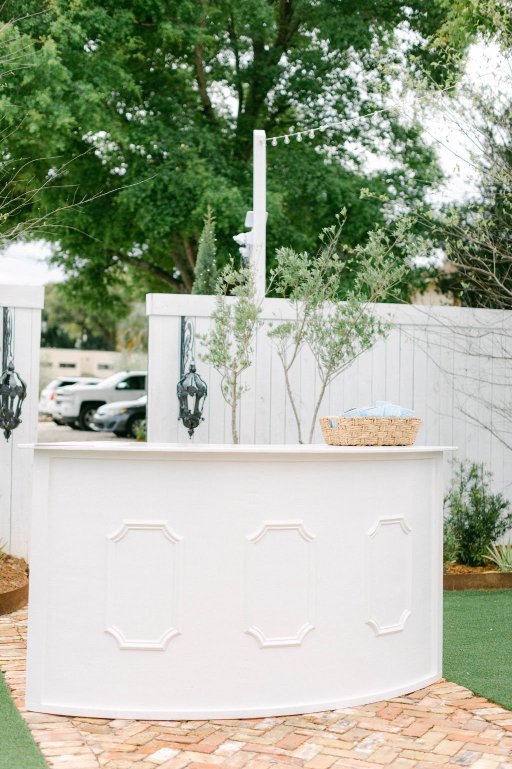 A white outdoor bar with decorative panels is set against a white wooden fence, with a basket of napkins on top. There are small trees and greenery behind the bar, in front of a large leafy tree, with a parking lot visible in the background.