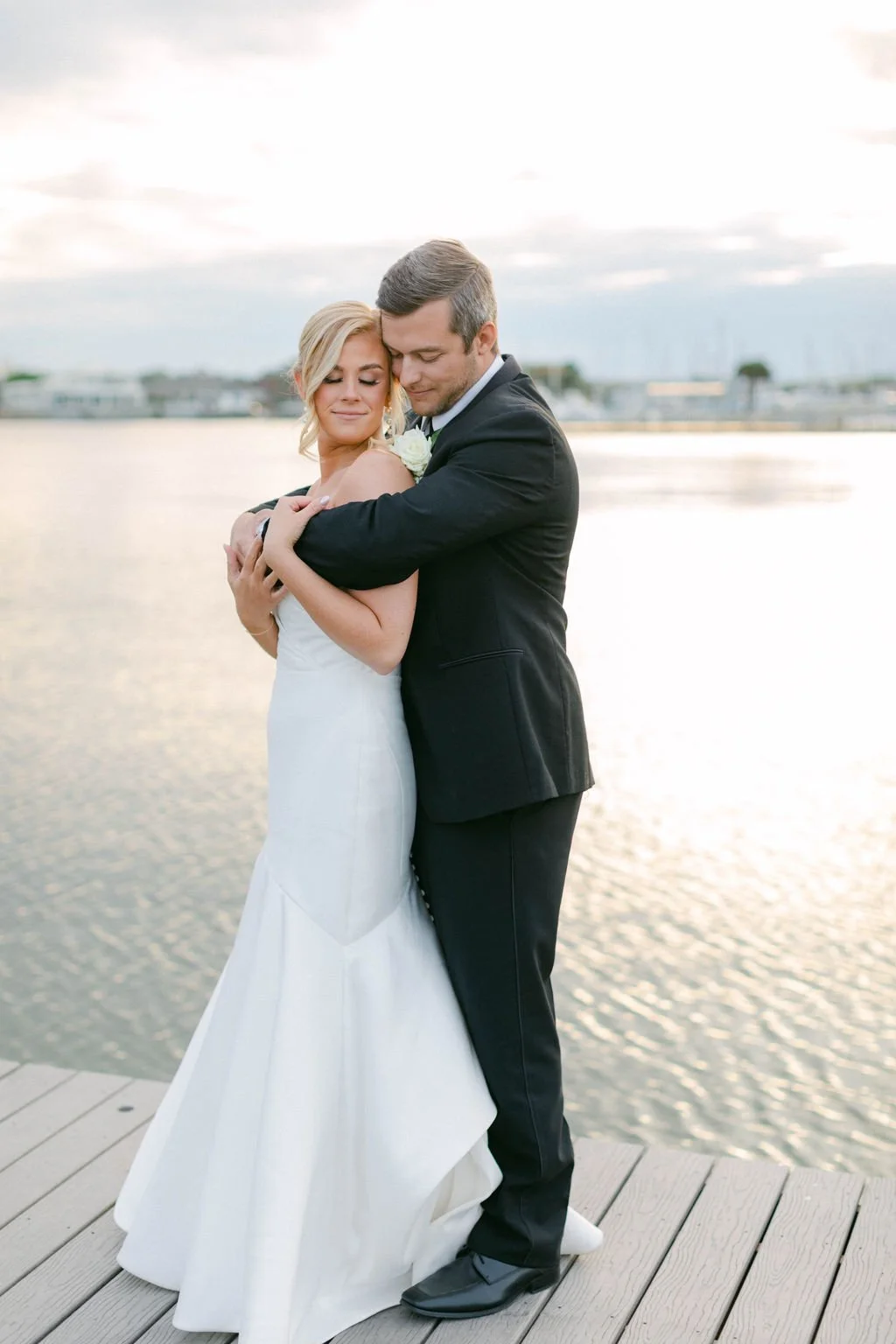 Bride in a white wedding gown and groom in a black tuxedo embrace on a wooden dock by the water, with a cloudy sky in the background.