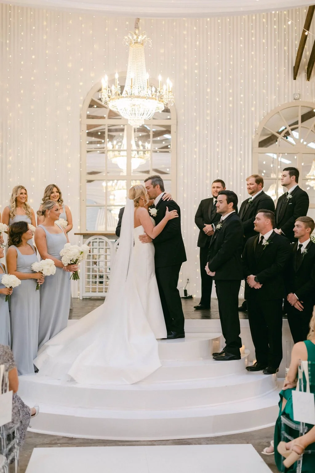A wedding ceremony with a bride and groom kissing on a white circular altar, surrounded by bridesmaids and groomsmen, in a decorated indoor venue with a chandelier and string lights.