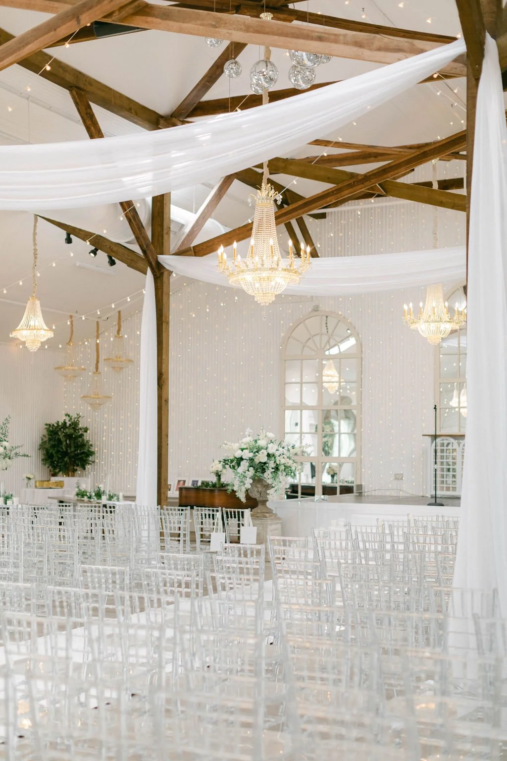 Elegant wedding venue decorated with white drapery, chandeliers, and string lights, featuring wooden beams, large mirror, and floral arrangements.