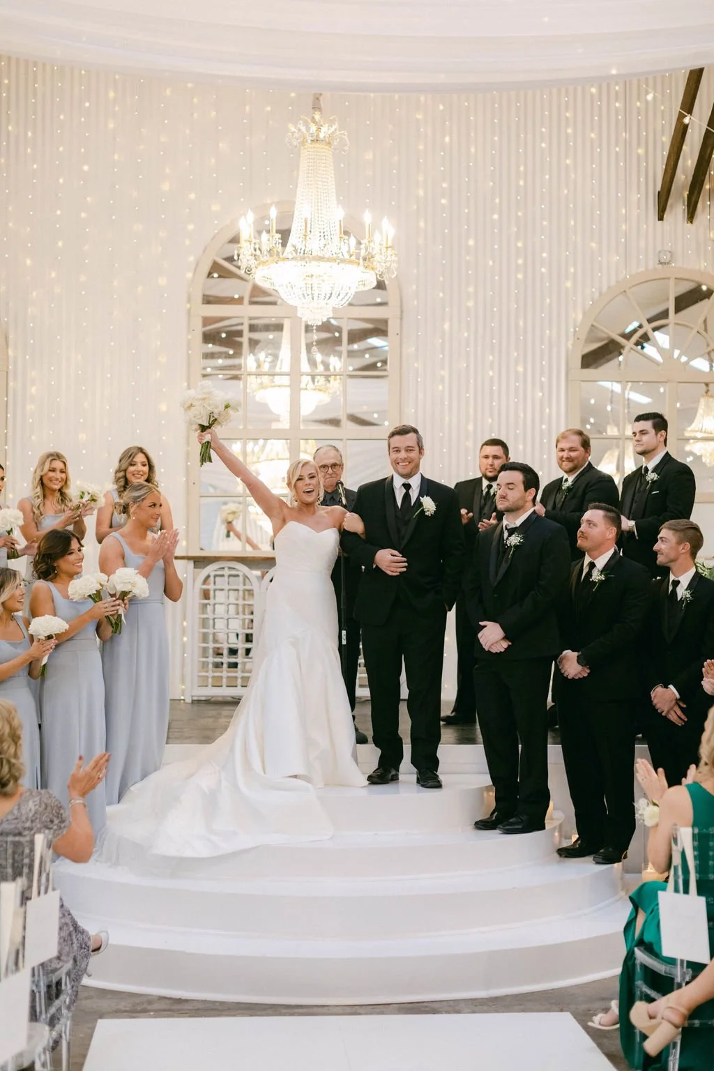 A wedding ceremony with the bride and groom on a stage, surrounded by bridesmaids and groomsmen, in an elegant decorated venue with chandeliers and string lights.