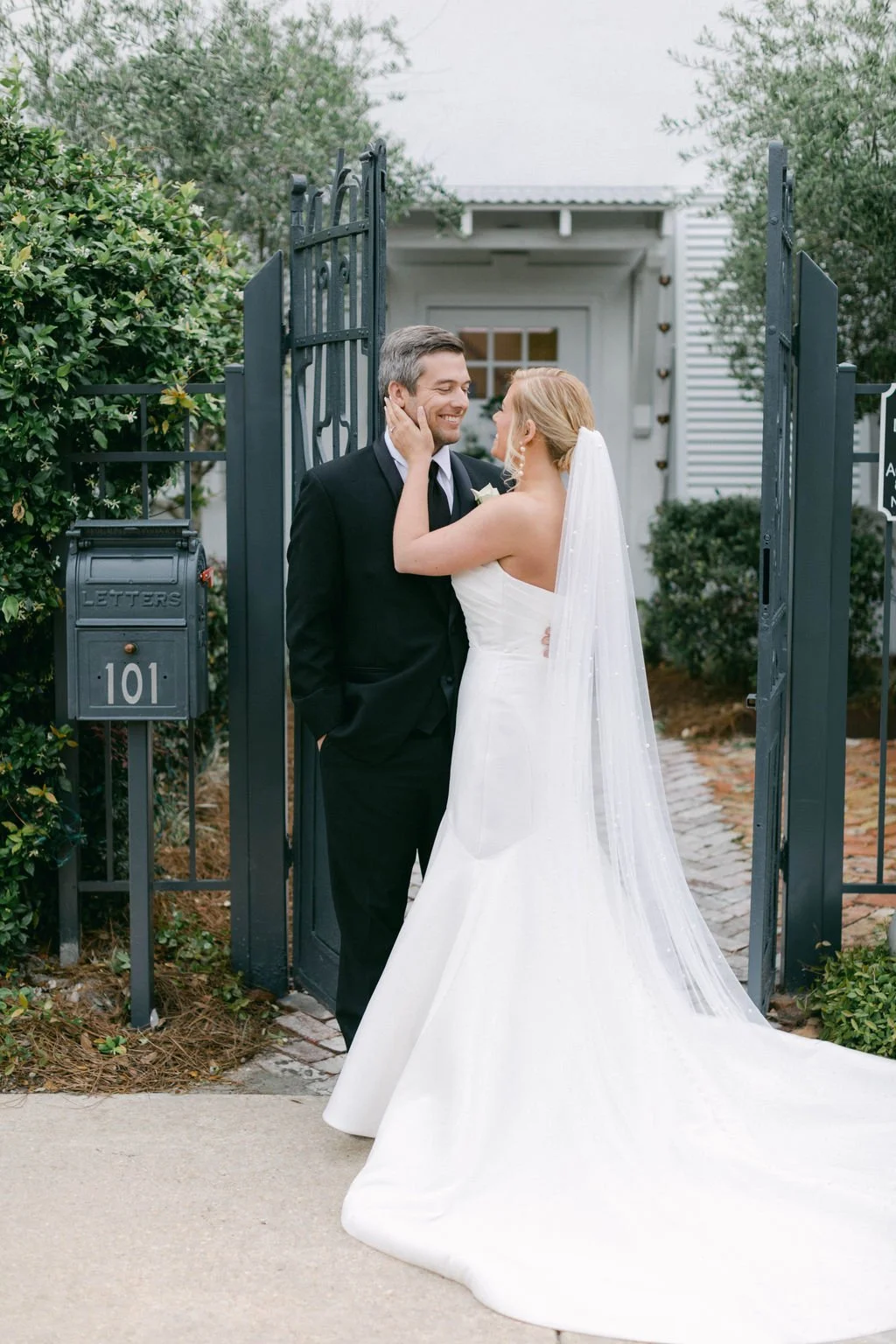 A bride and groom embrace in front of a black iron gate, with the bride in a strapless white wedding gown and veil, and the groom in a black tuxedo.