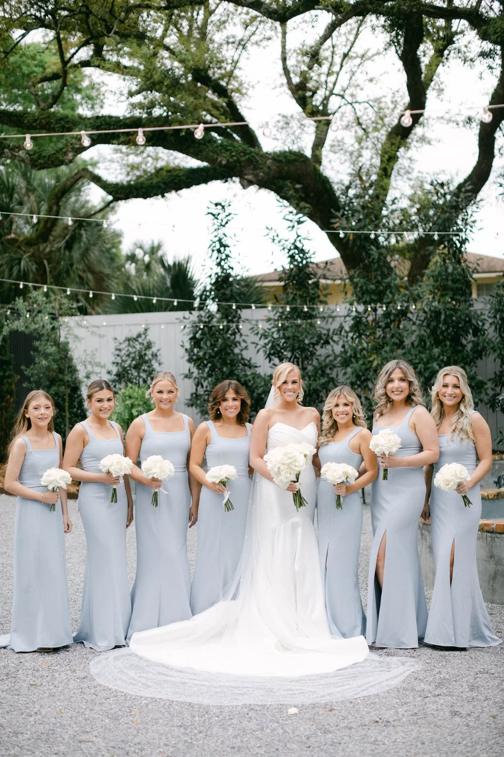 A bride and seven bridesmaids pose outdoors on a gravel surface, with the bride in a white wedding gown and the bridesmaids in matching light gray dresses, each holding a bouquet of white flowers. Large trees and string lights are visible in the back