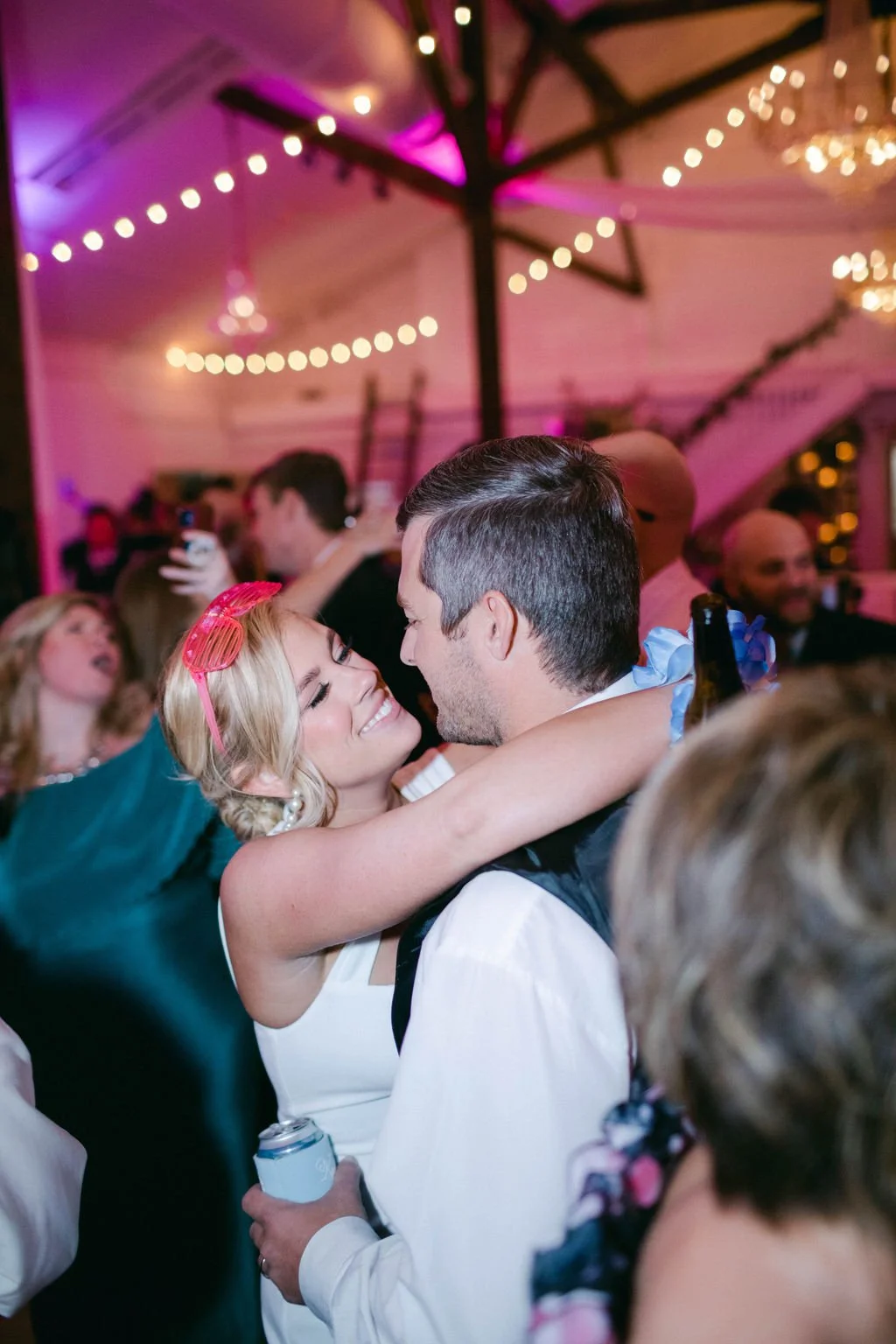 A couple is embracing and smiling at each other at a lively party or wedding reception with colorful string lights and people dancing in the background.