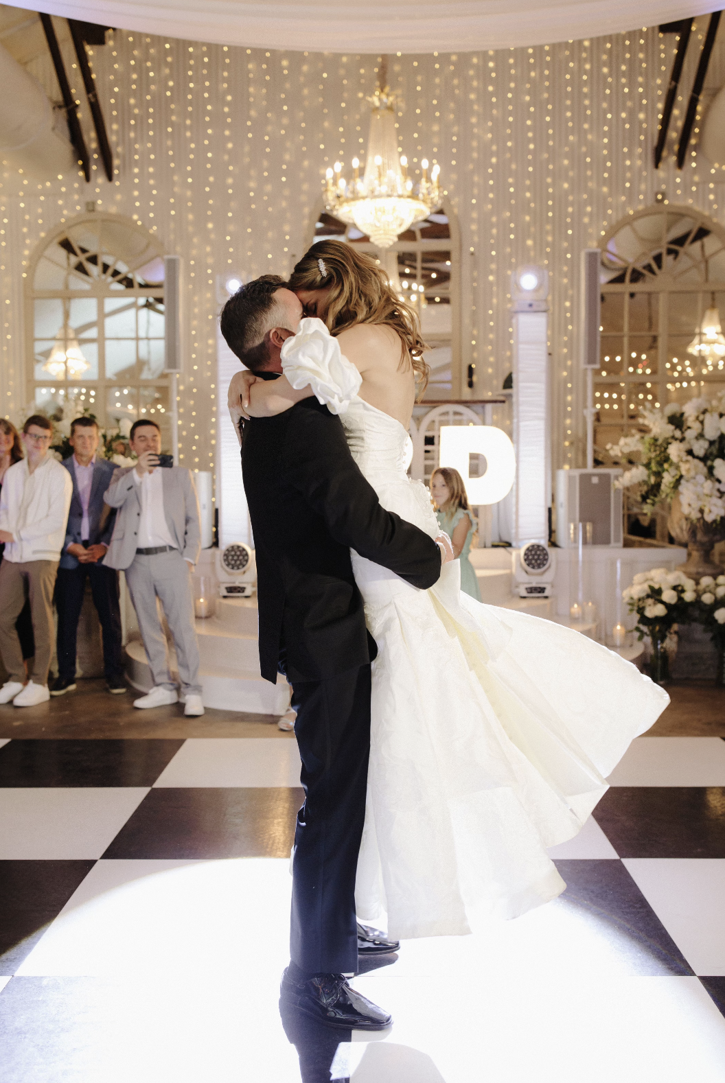 Couple dancing at a wedding reception, with the man lifting the woman in a white wedding dress, amid guests and fairy lights in an elegant decorated venue.