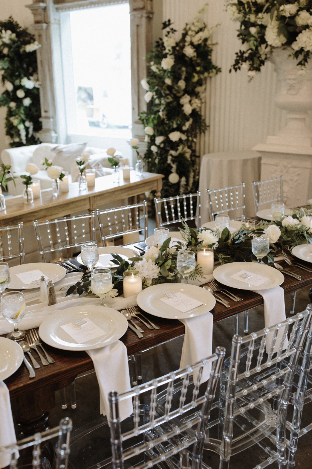 Elegant wedding reception table set with white dinnerware, silverware, glassware with lemon slices, white candles, and a green floral arrangement on a dark wooden table with clear acrylic chairs, decorated with white fabric draped over the chairs, in