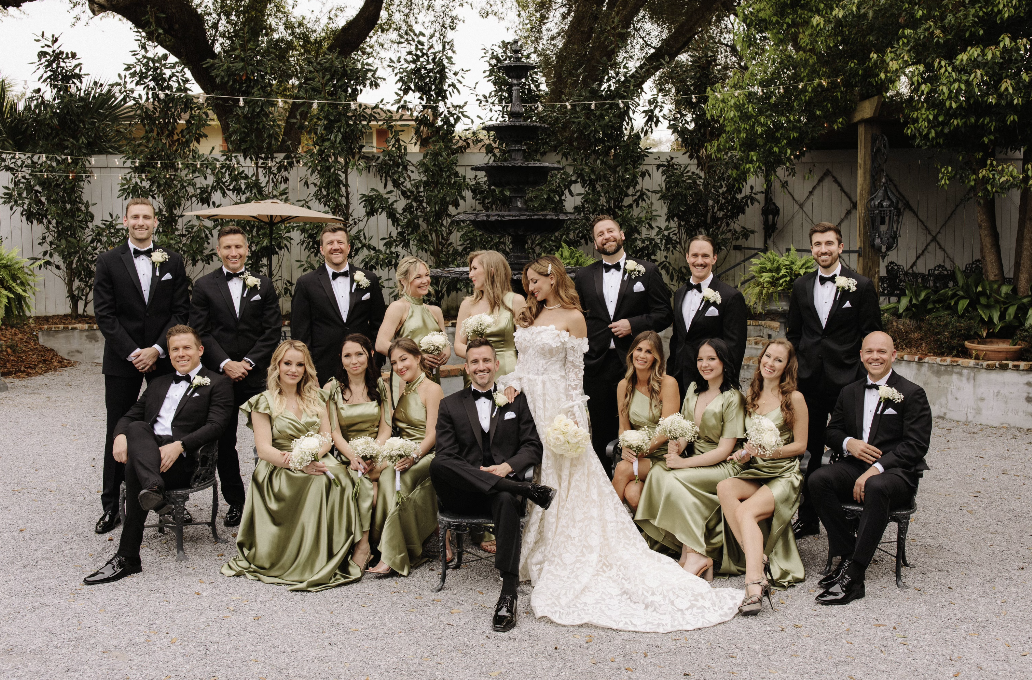 A wedding party of 20 people, including a bride in a white lace gown with a bouquet, groom in a black tuxedo, and 18 bridesmaids and groomsmen, outdoors in a garden setting with trees, plants, and a fountain in the background.