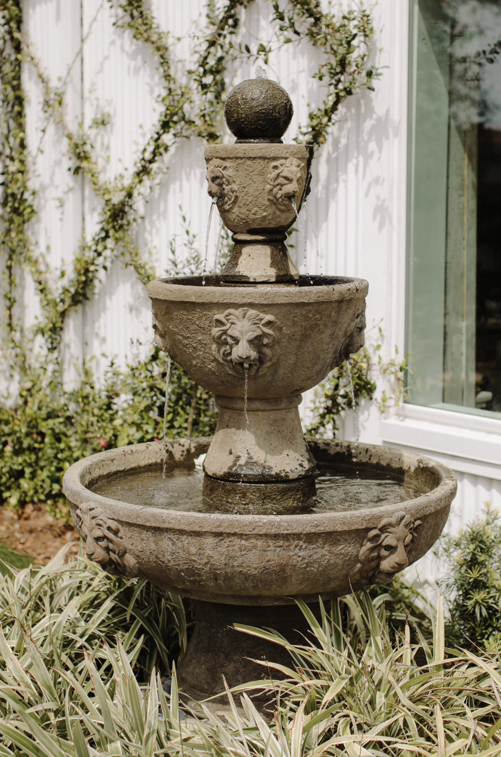 A multi-tiered stone fountain featuring lion head sculptures with water flowing from their mouths, situated outdoors with greenery and a white fence background.