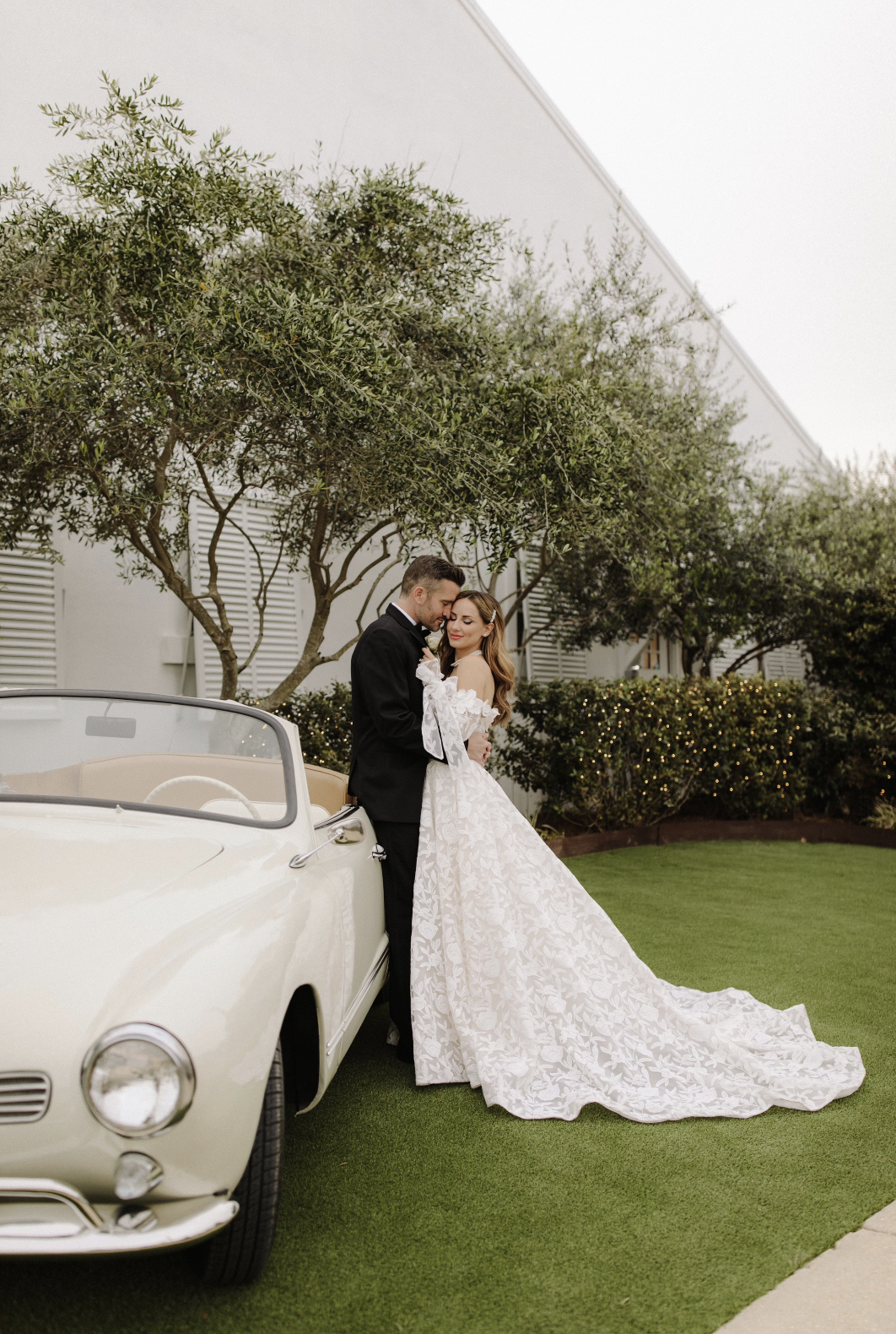 A bride and groom in wedding attire embracing next to a vintage white convertible car on a manicured lawn with trees and bushes in the background of a Pensacola Wedding Venue.