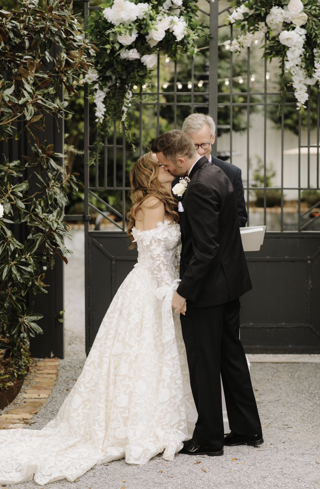 A Pensacola bride and groom share a kiss during their wedding ceremony, standing in front of a black gate decorated with white flowers, with an officiant behind them.