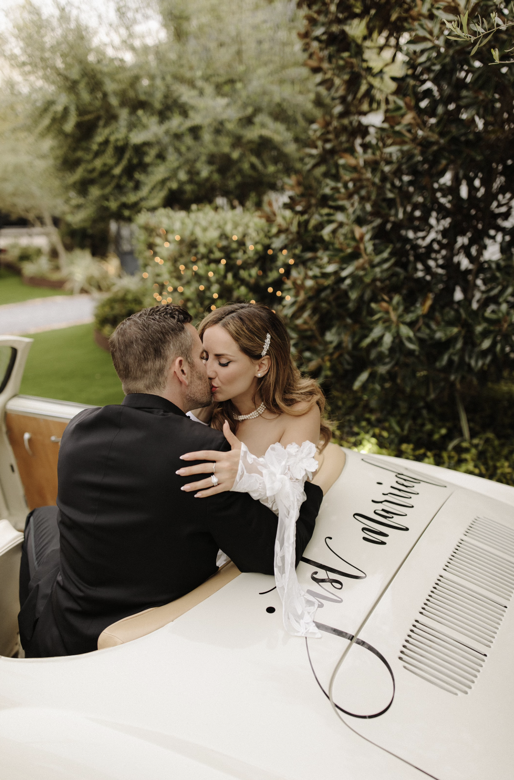 Couple in wedding attire sharing a kiss while sitting in a vintage car with wedding signs on the hood, outdoors in a garden setting.