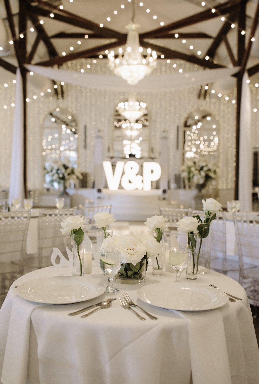 Wedding reception table with white roses, glassware, and silverware, decorated with illuminated 'V&P' sign in the background, hanging string lights, draped fabric, and chandeliers in an elegant indoor venue.