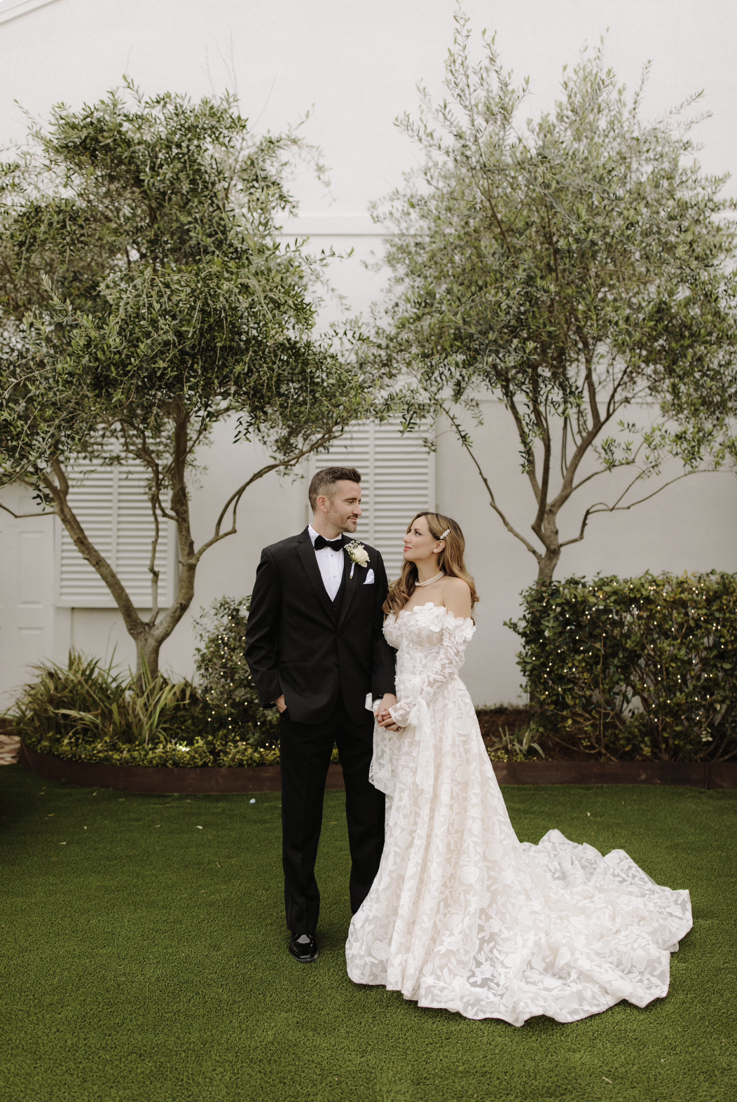 A Pensacola bride and groom holding hands outdoors on a wedding day, surrounded by trees and greenery.