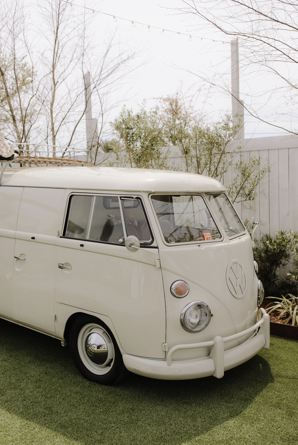 A vintage cream-colored Volkswagen Type 2 van parked on artificial grass in a backyard, surrounded by plants and trees with a white fence and string lights overhead.