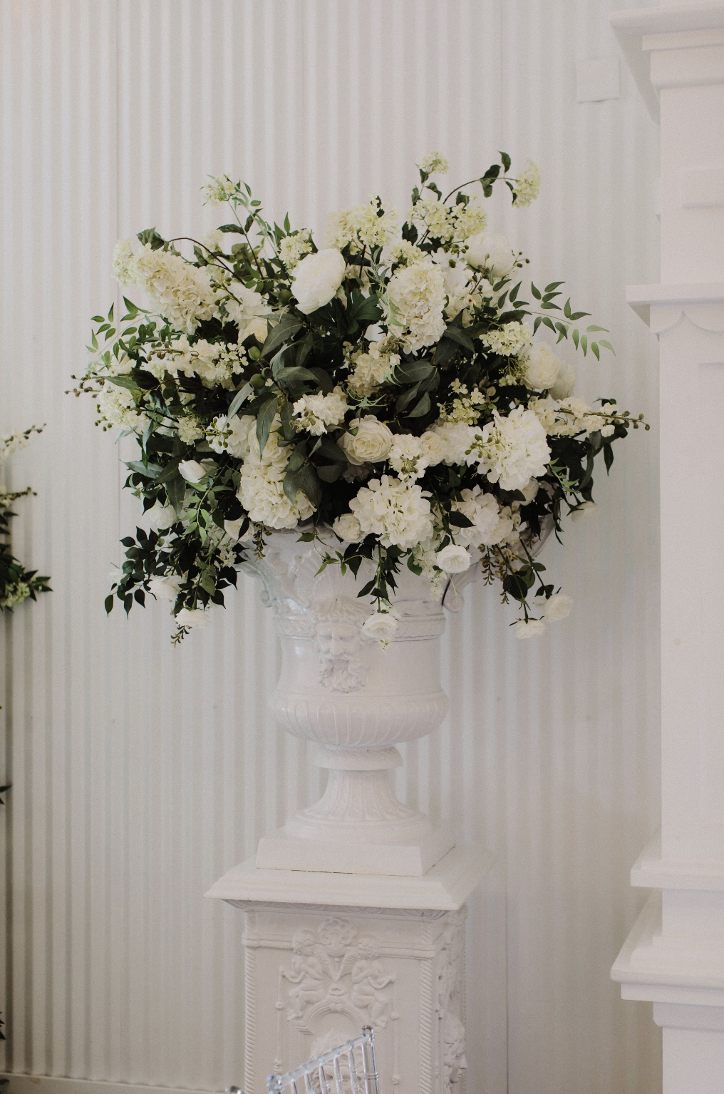 A large white floral arrangement in an ornate white vase on a pedestal, featuring white roses, hydrangeas, and greenery.