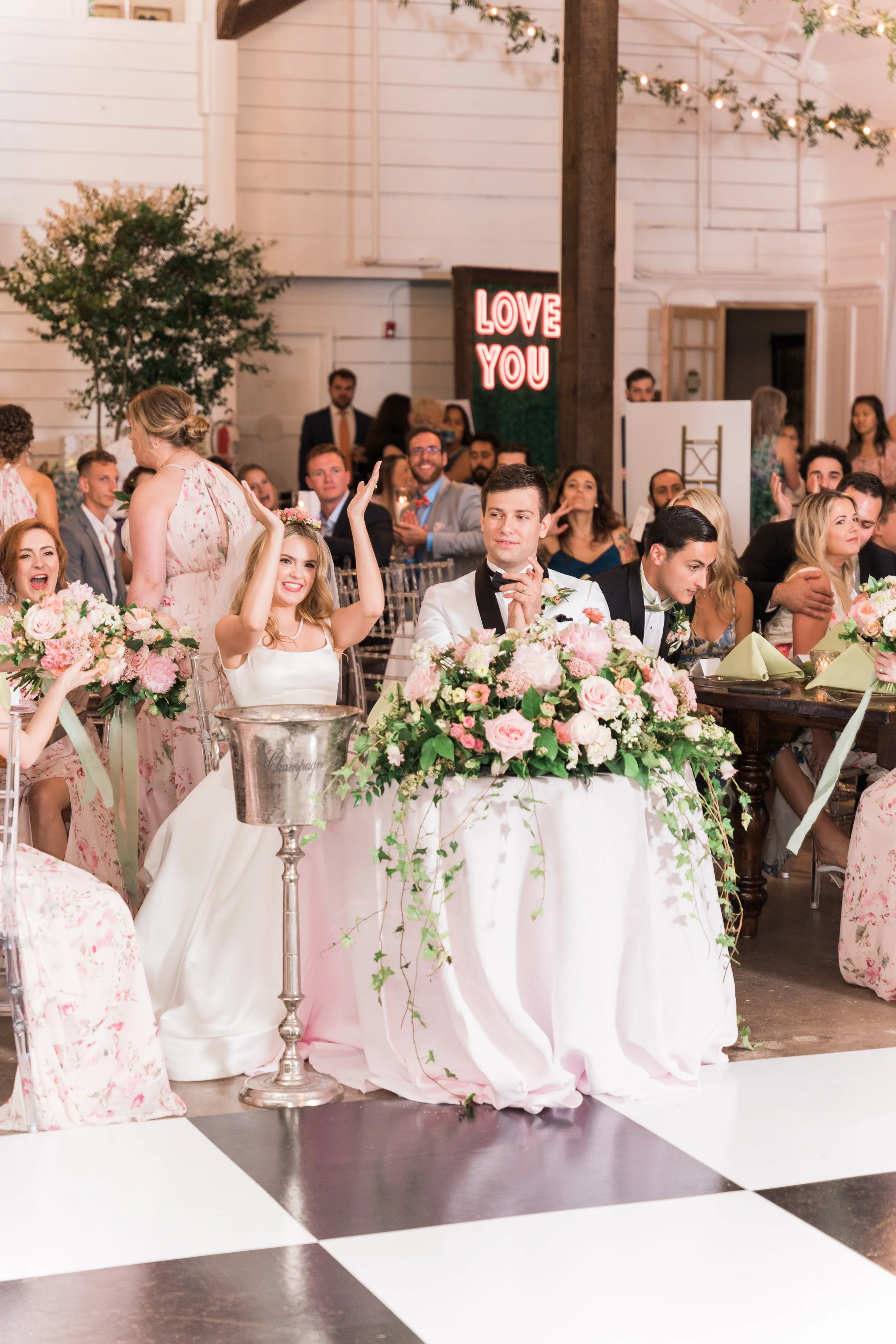 Pensacola Wedding reception with guests seated around tables decorated with pink and white flowers, a bride and groom sitting at the head table, and a neon sign in the background that says 'LOVE YOU'.