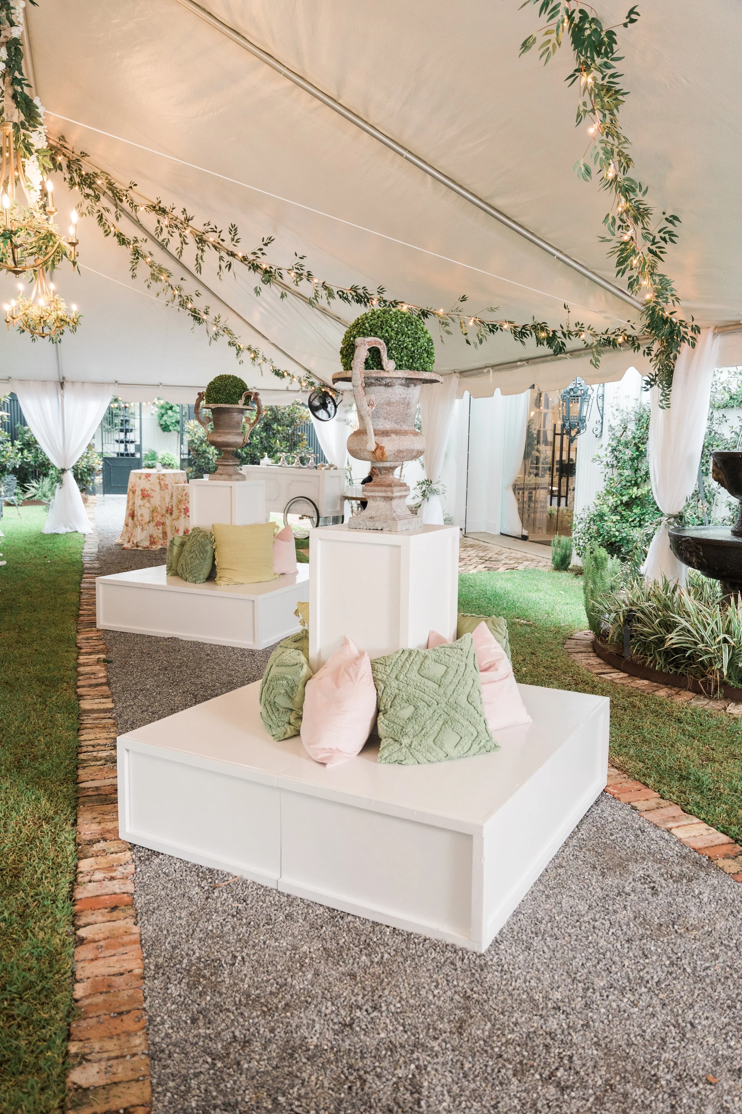 Elegant outdoor tent decorated with greenery and string lights, featuring white platforms with pastel-colored pillows, decorative urns, and lush plants for an event or wedding.