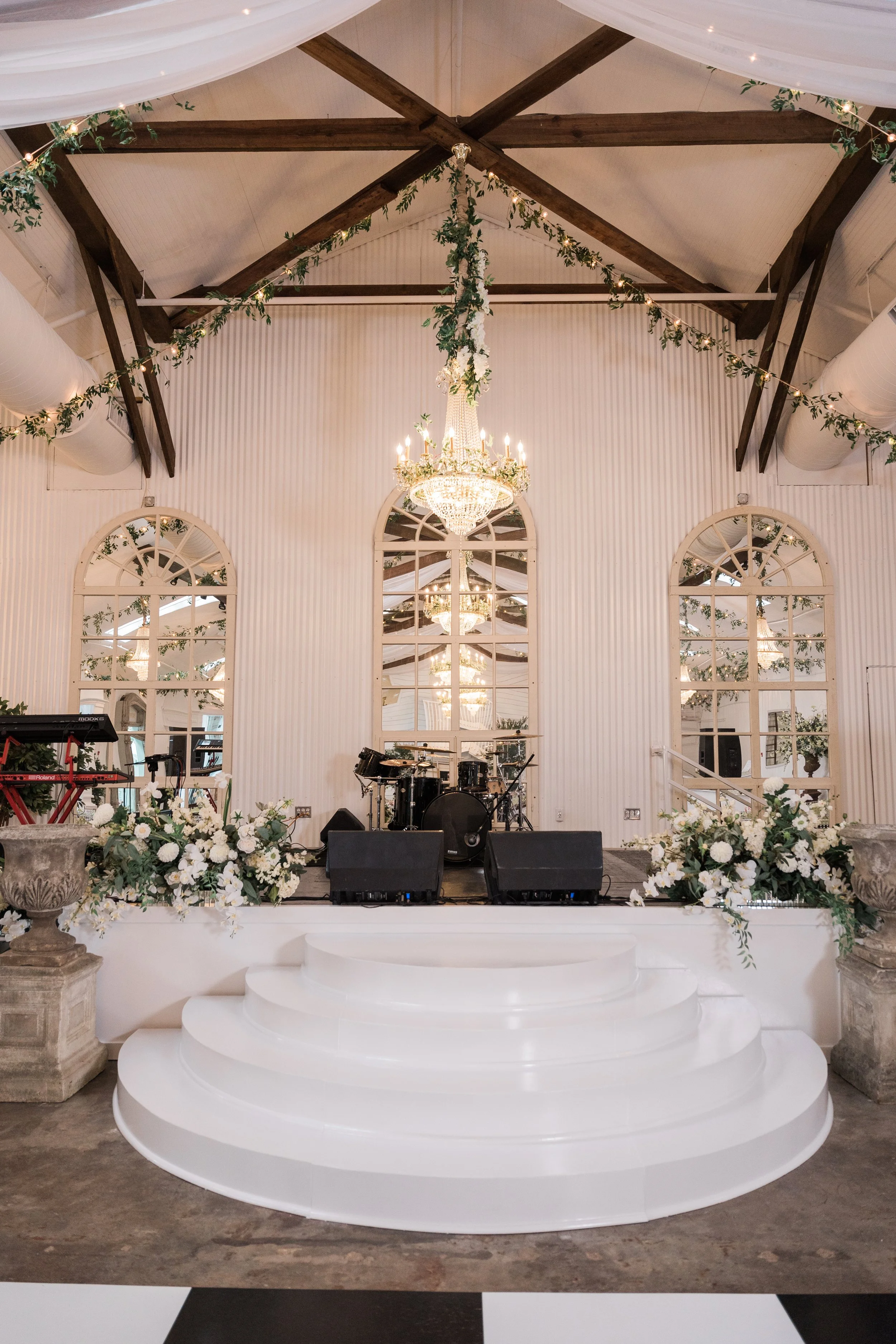 An elegantly decorated indoor event space with a small stage featuring musical instruments, surrounded by floral arrangements, large mirrors, chandeliers, and string lights with greenery, under a high ceiling with wooden beams and draped white fabric