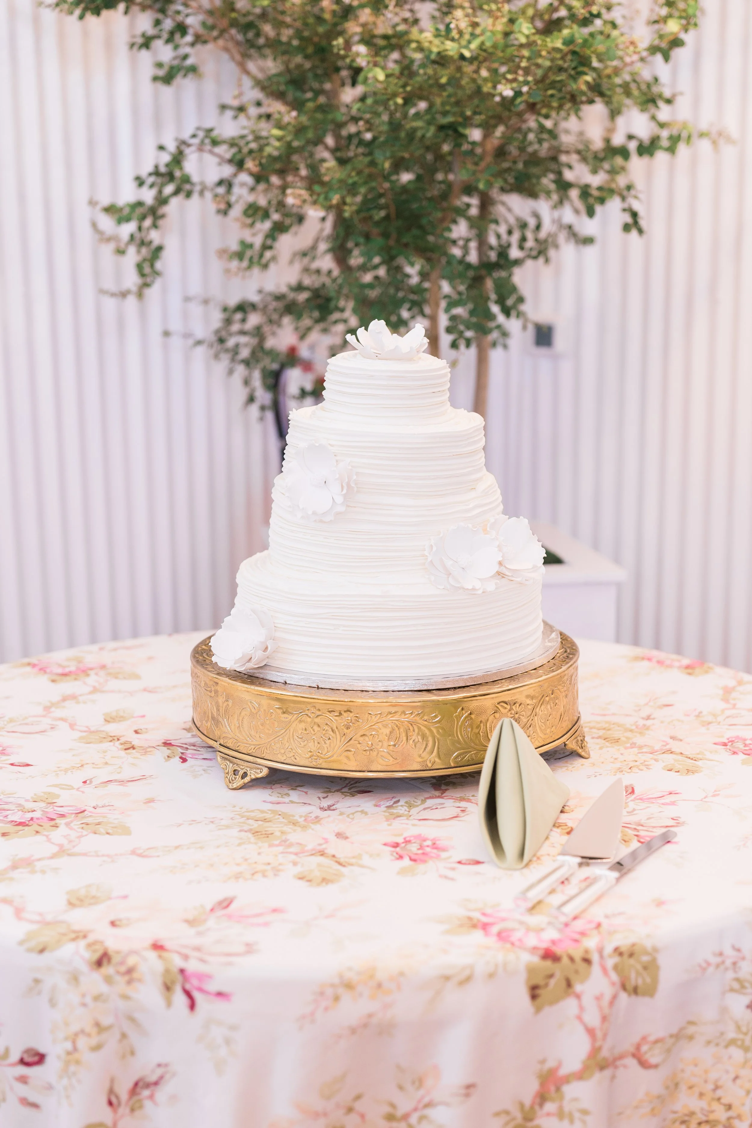 Three-tiered white wedding cake with floral decorations on a gold stand, set on a floral tablecloth with a green plant in the background.