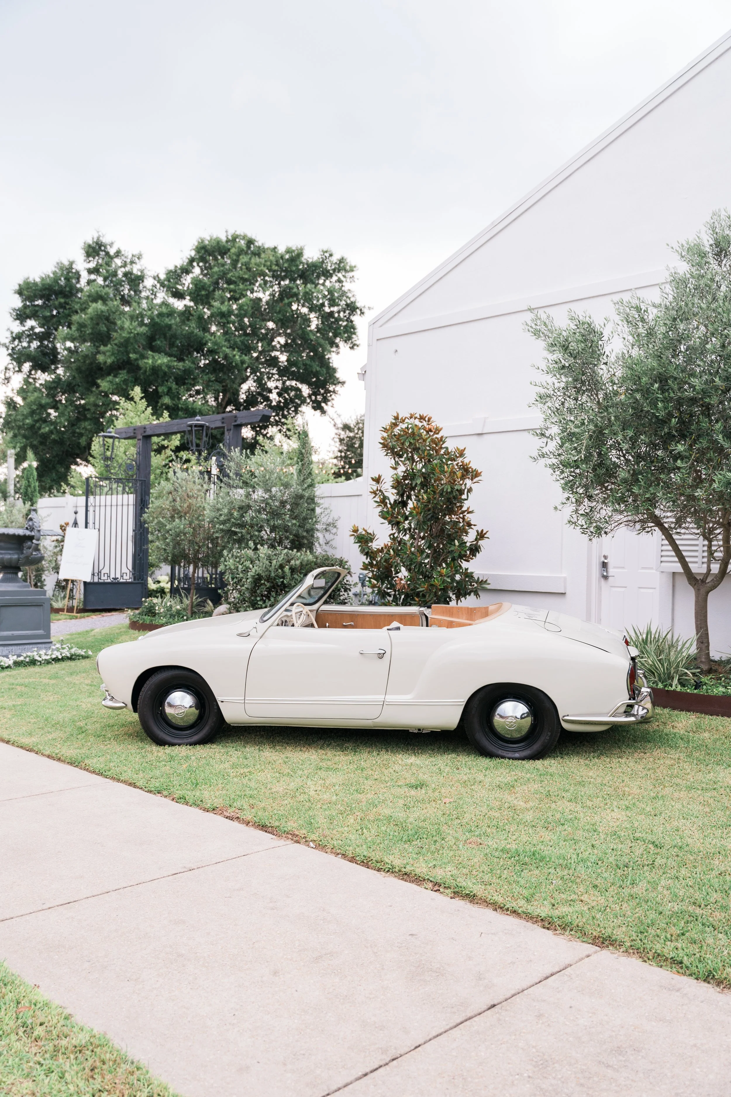 A vintage white convertible car parked on a well-manicured lawn beside a white building with trees and bushes in the background of a Pensacola Wedding Venue.