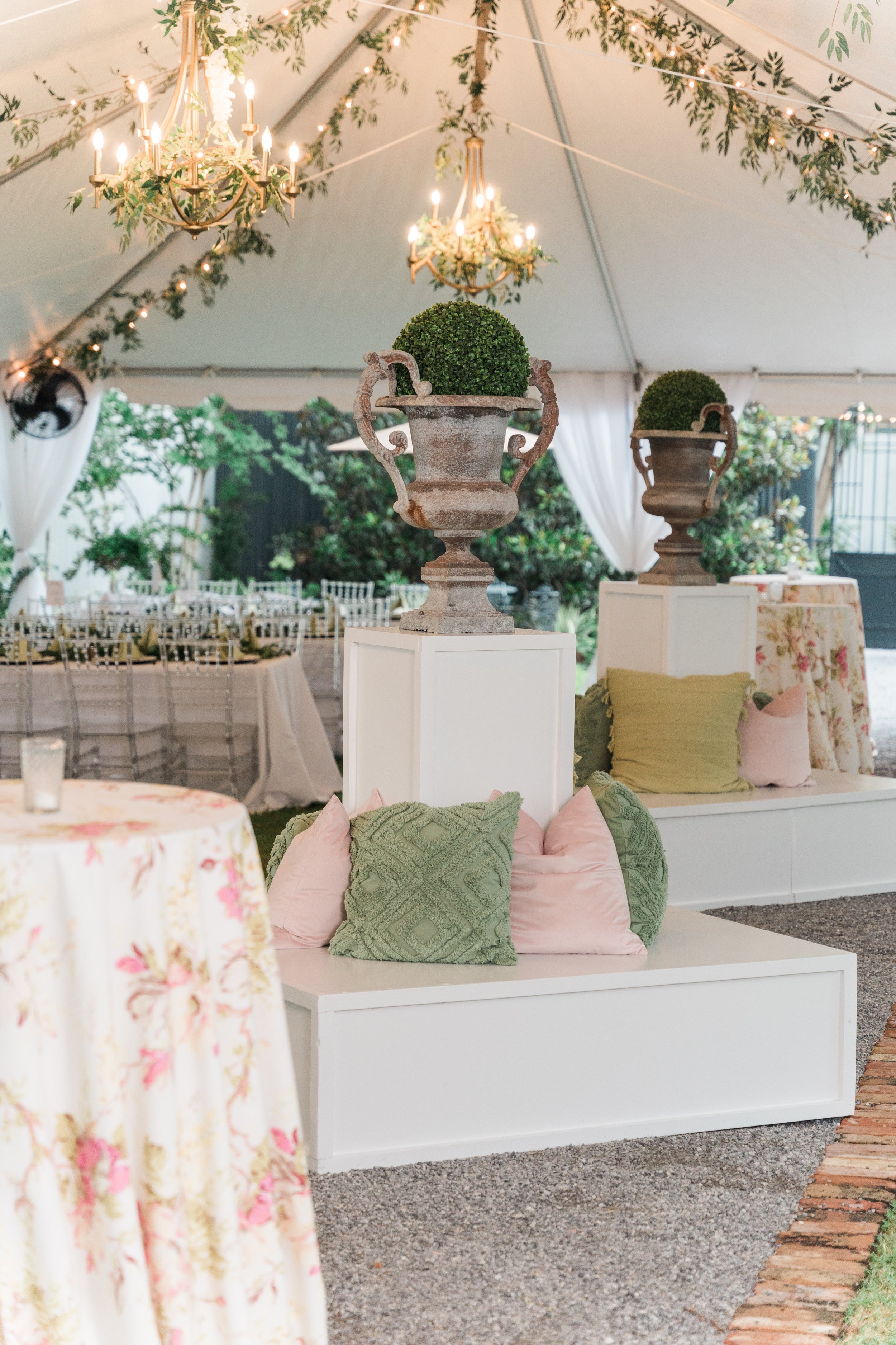Decorative setup inside a white event tent with chandeliers, greenery, and elegant furniture, including planters with topiary and decorated pillows.