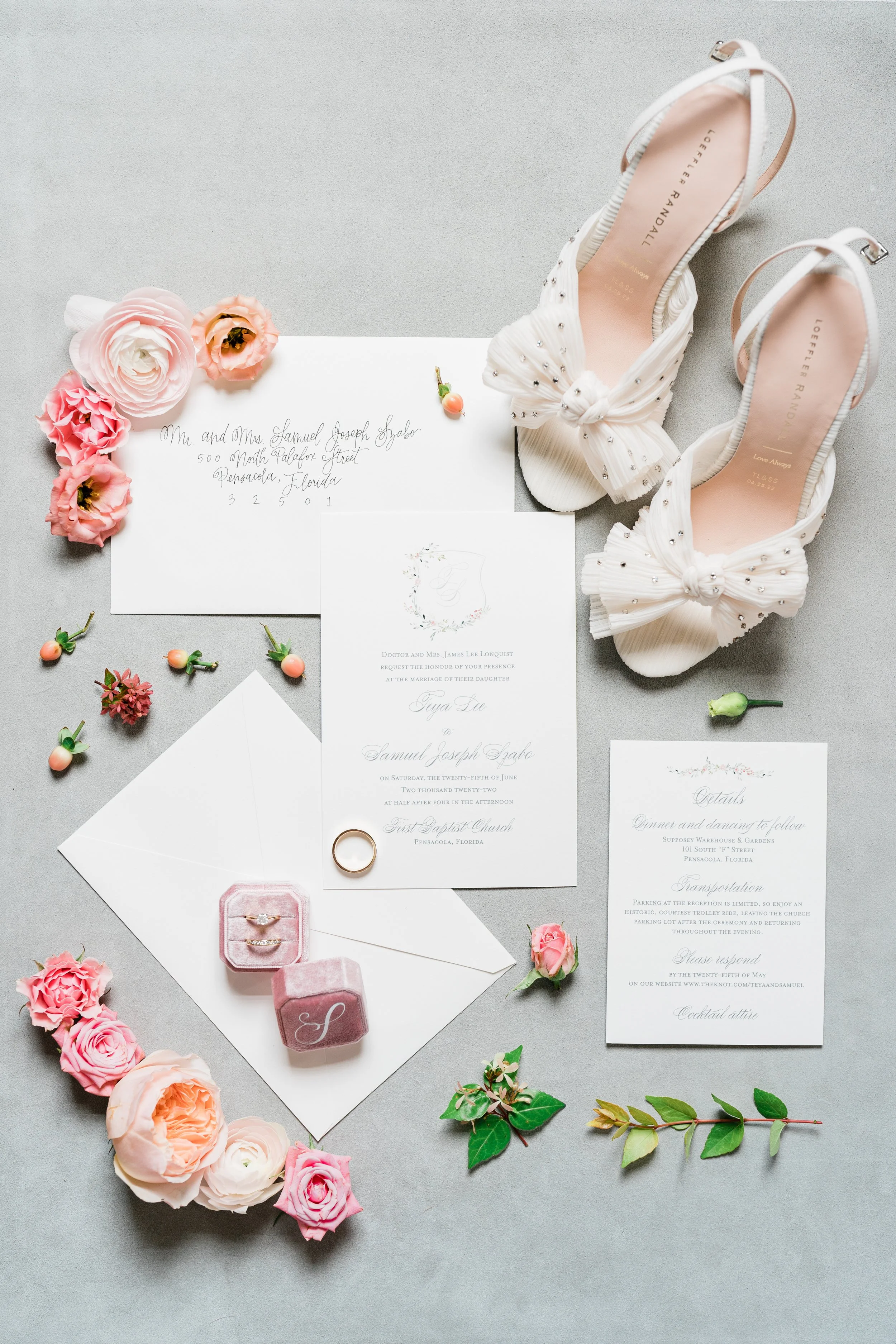 Wedding invitation, pink rose blossoms, a gold wedding ring, a pair of white high-heeled shoes with bows, and decorative greenery arranged on a gray surface.