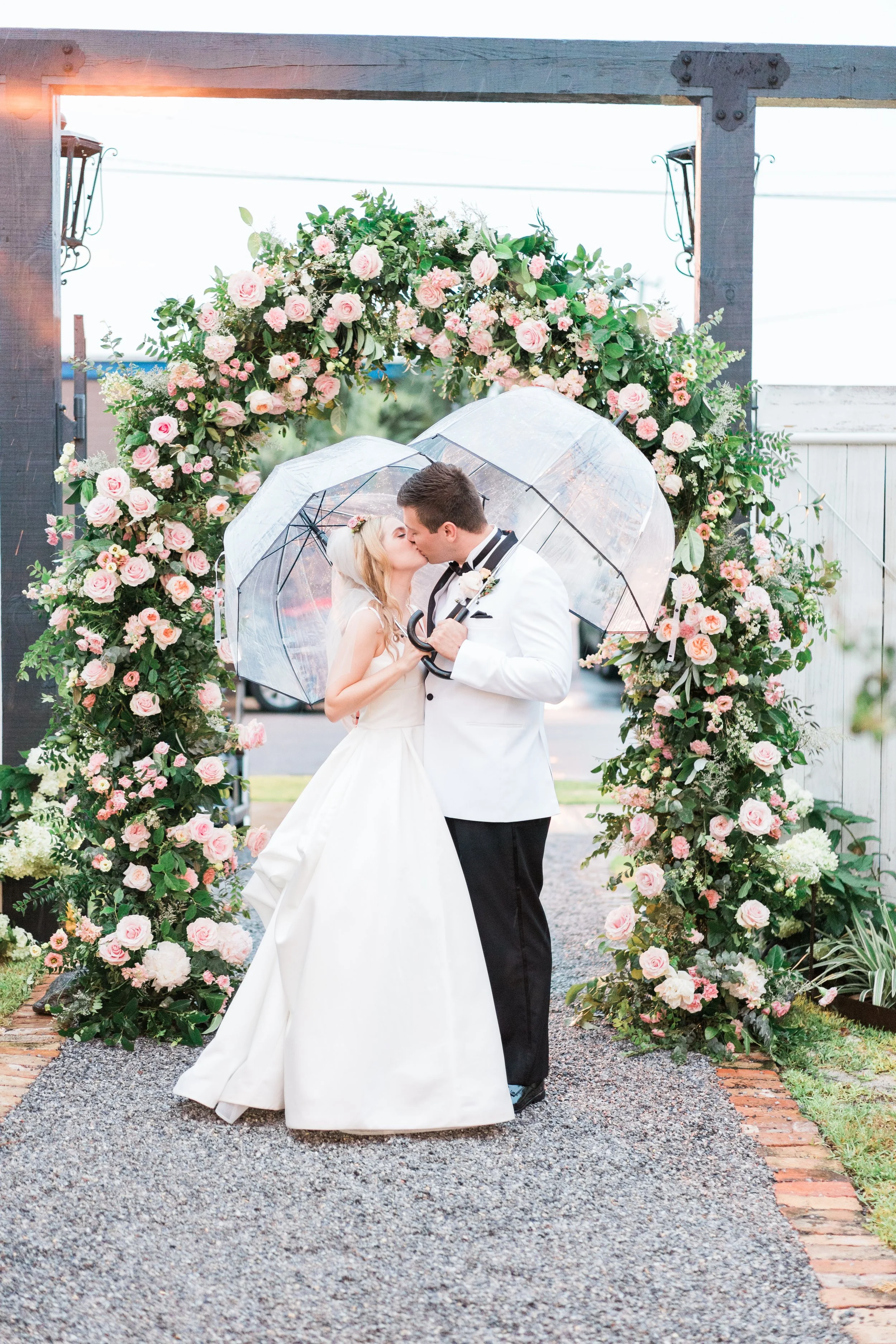 A newlywed couple shares a kiss under clear umbrellas, standing in front of a floral arch decorated with pink and white roses, on a gravel pathway.
