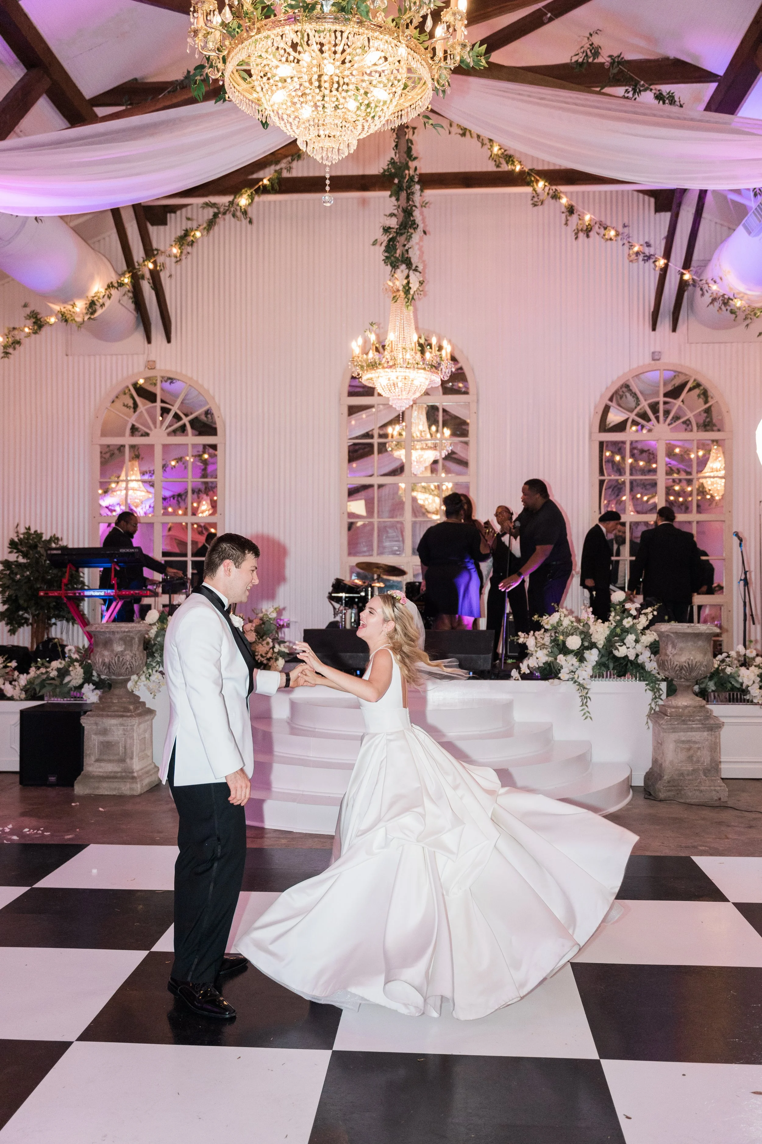 Bride and groom dancing at their wedding reception, with band playing in background, decorated with elegant chandeliers, floral arrangements, and draped fabric ceiling.