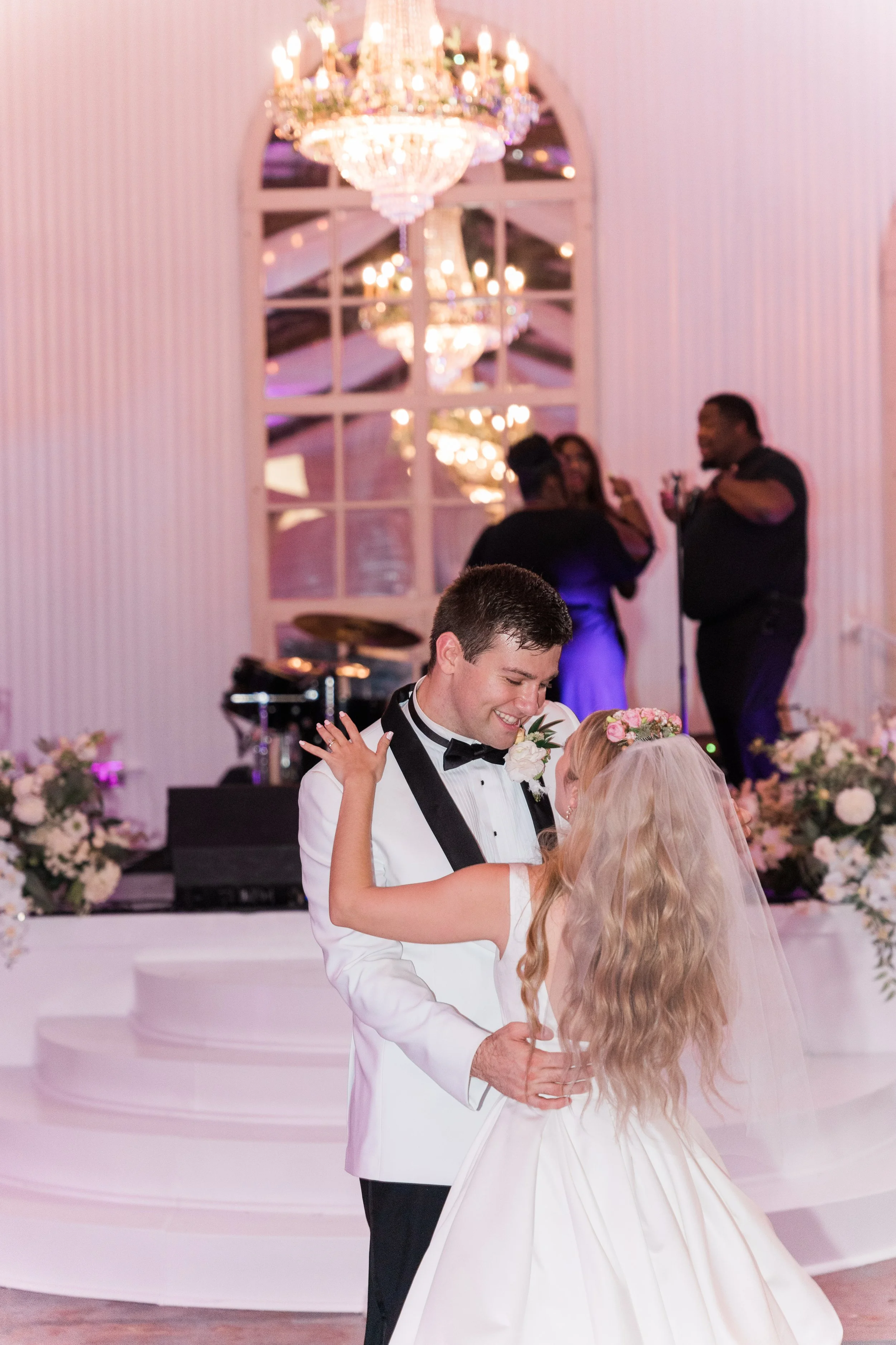 A bride and groom dancing at a wedding reception, with a chandelier hanging above and other guests in the background.