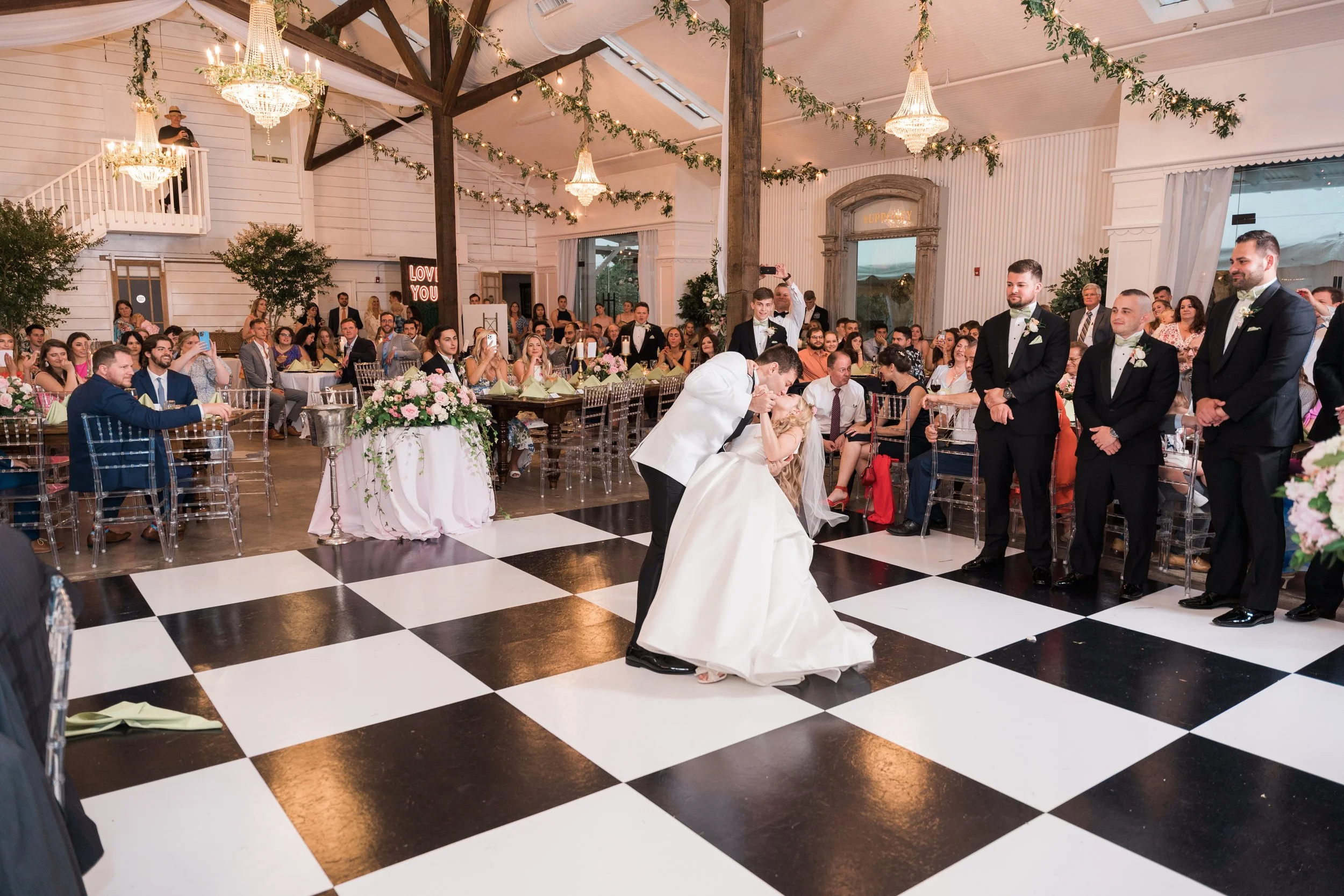 A wedding reception scene in a decorated hall with a couple sharing a dance on a checkered black and white floor. The bride is in a white gown, and the groom is in a tuxedo. Guests are seated at decorated tables with pink floral arrangements, watchin