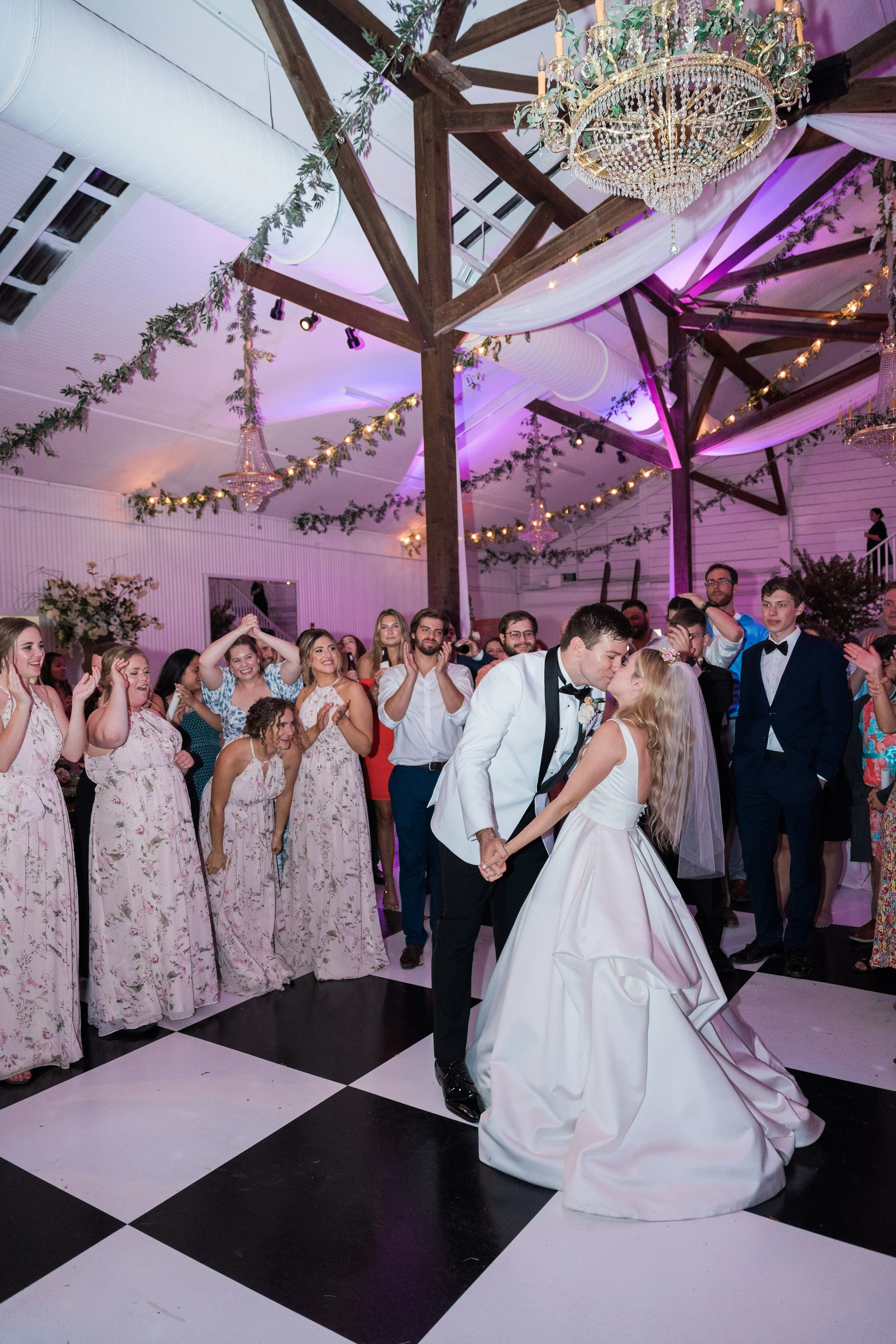 A bride and groom are dancing and kissing at their wedding reception with guests surrounding them, in a decorated venue with chandeliers, string lights, and draped fabric.