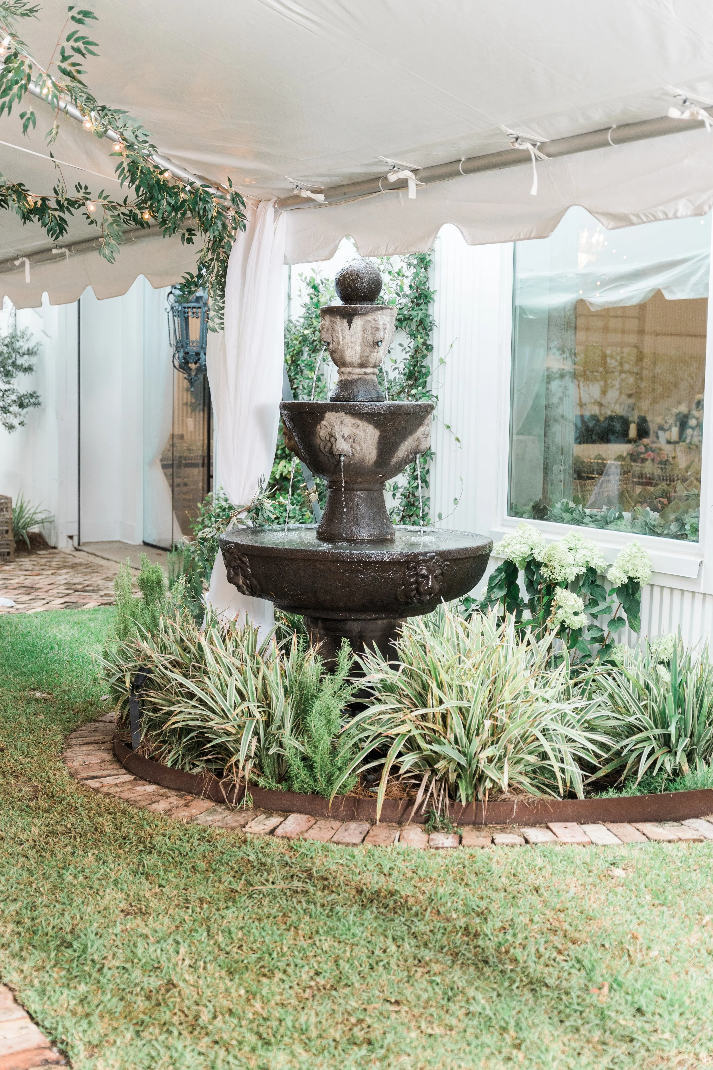 A decorative outdoor fountain with a lion face spout, surrounded by green plants, under a white canopy tent with string lights.