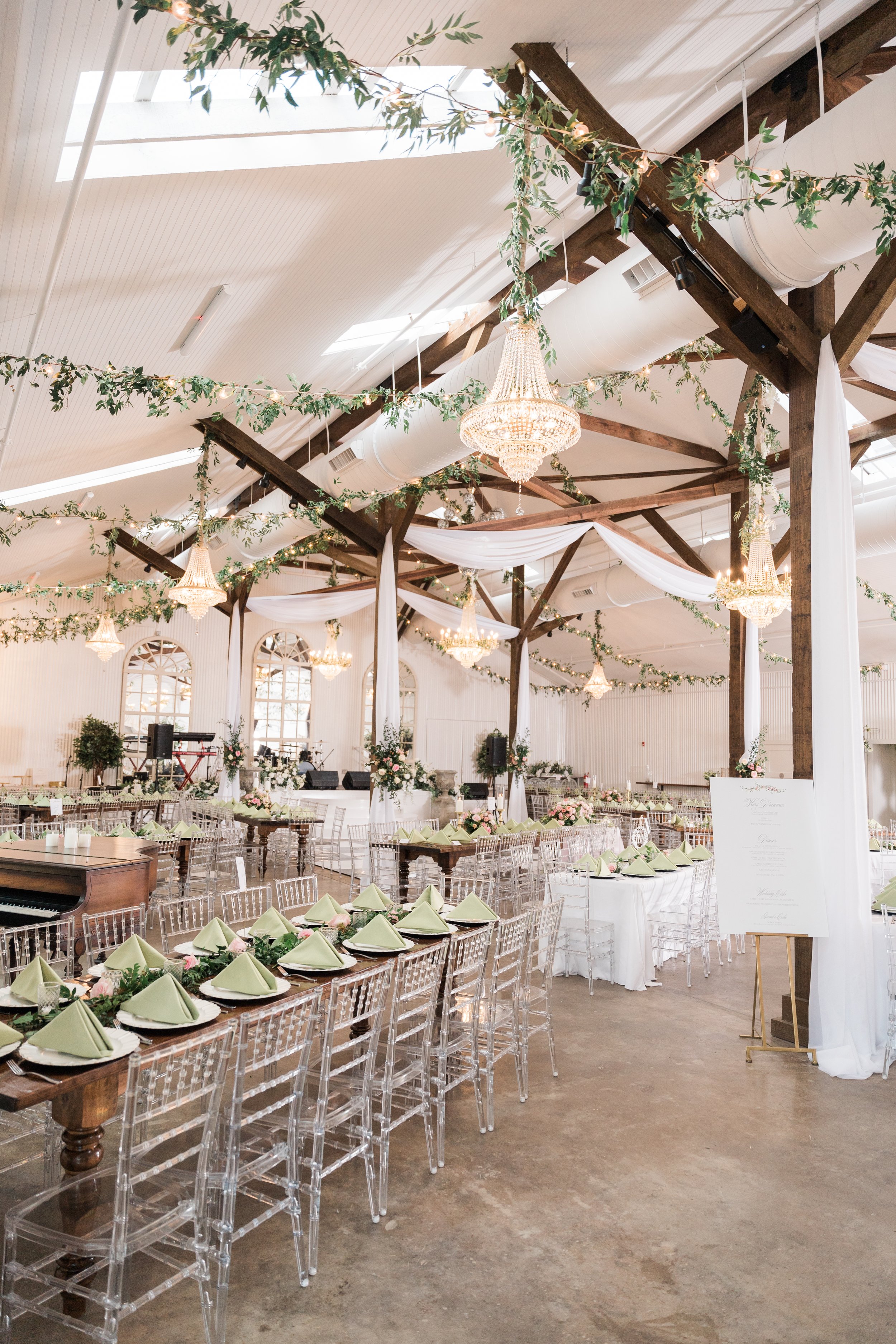 Elegant event hall decorated with chandeliers, draped white fabric, and green foliage. Long tables with transparent chairs are set with green napkins and floral centerpieces. The ceiling has skylights and exposed wooden beams.