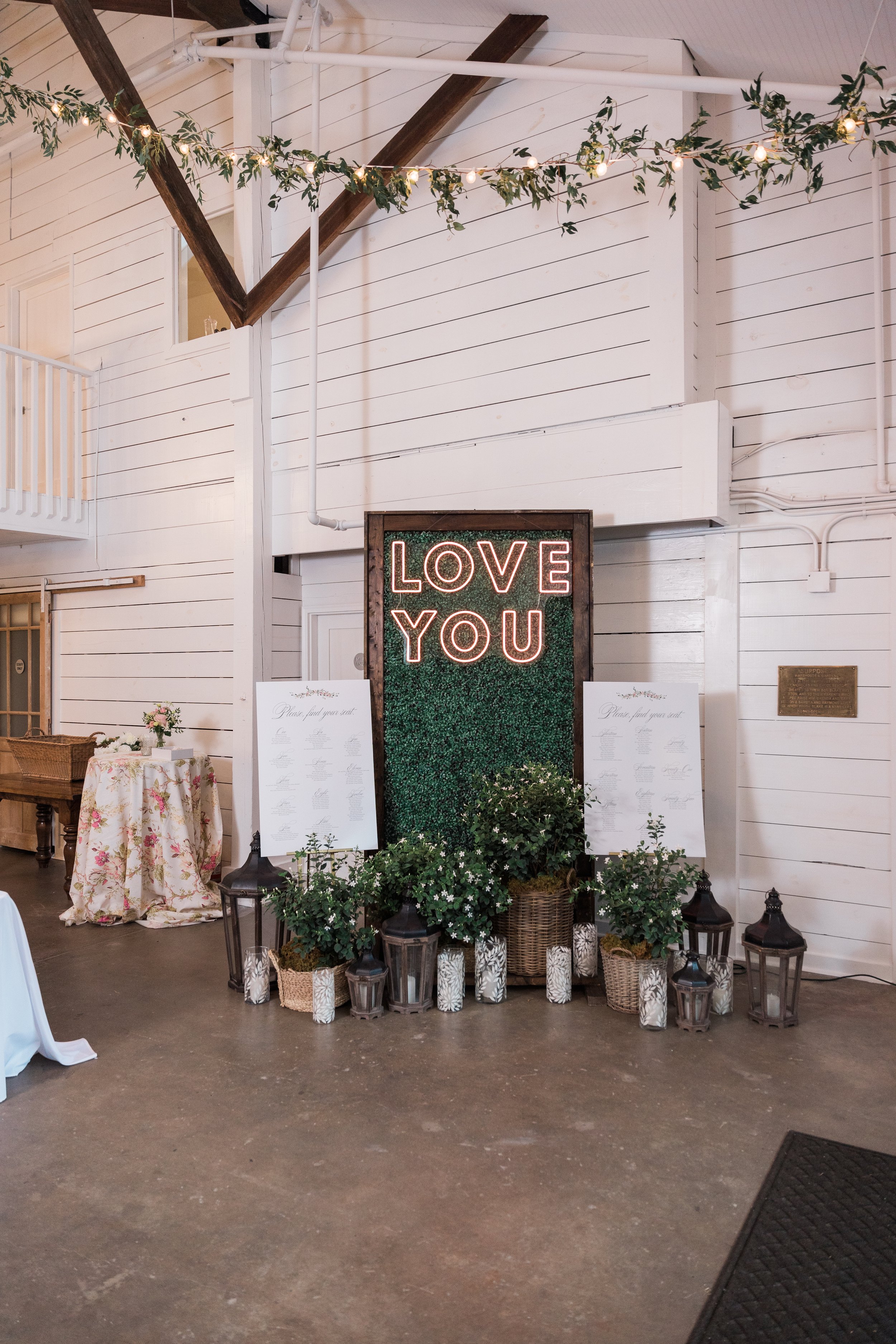 A decorated indoor event space with a neon sign saying 'LOVE YOU' above a green foliage wall, surrounded by potted plants, lanterns, and seating charts, with floral and string lights decorations.
