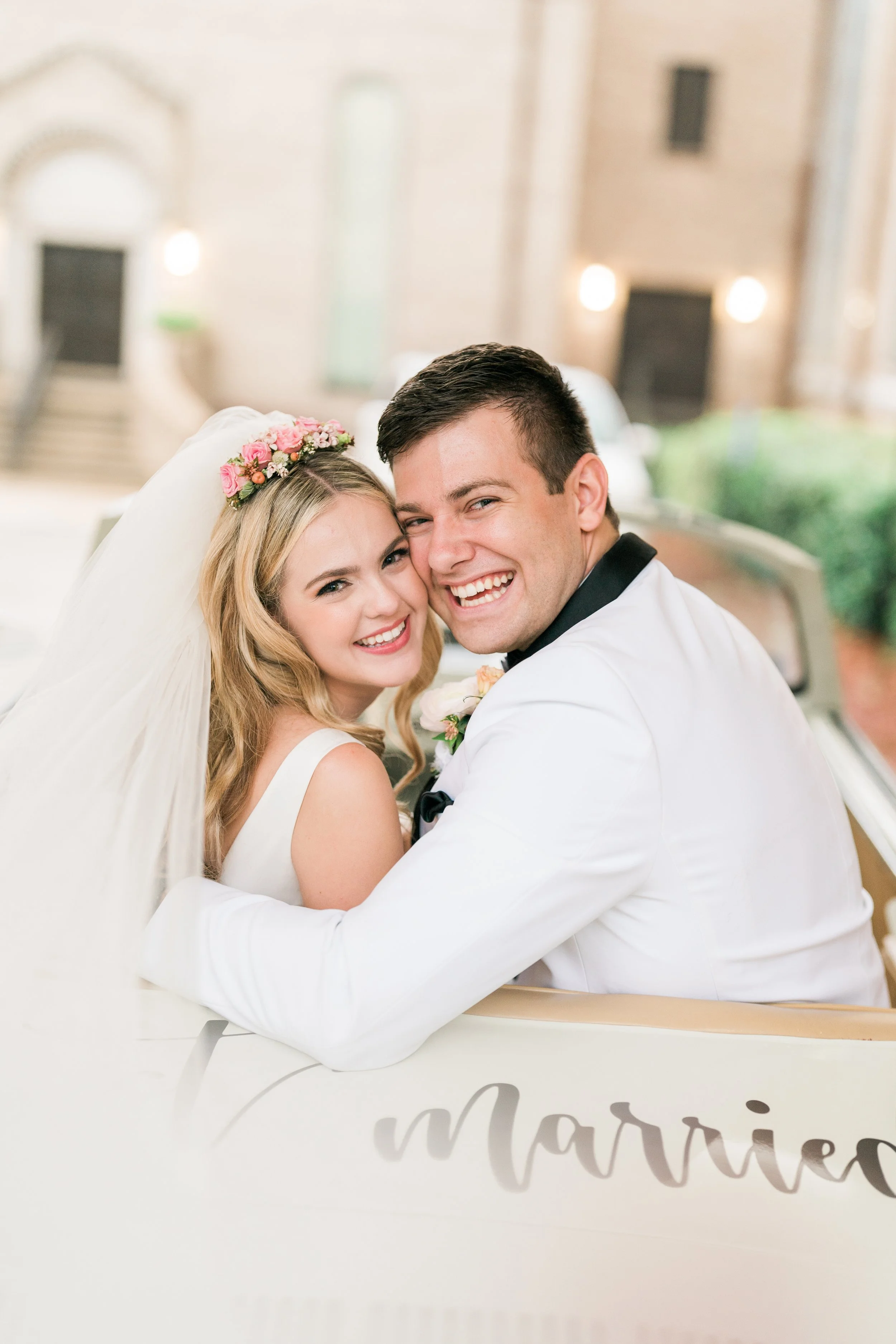 A smiling bride and groom embracing on a white vintage car with the word 'married' written on it, celebrating their wedding in Pensacola.