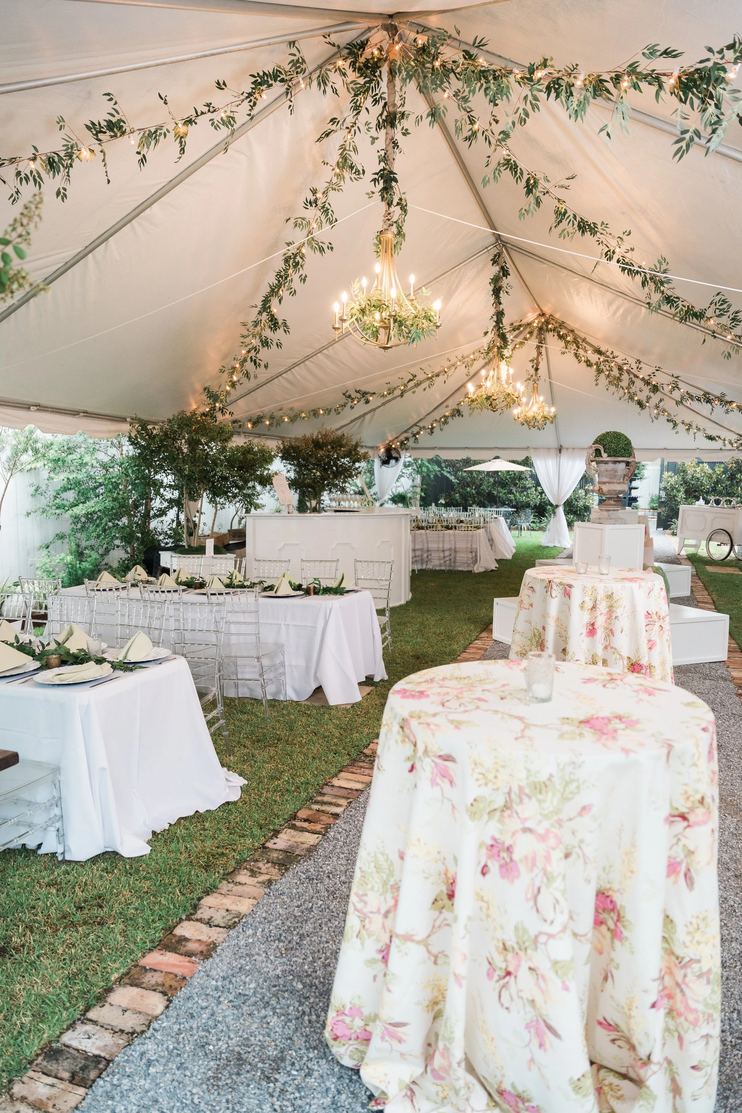 Elegant outdoor event setup under a tent with decorated chandeliers, greenery, and tables with white tablecloths and floral arrangements.