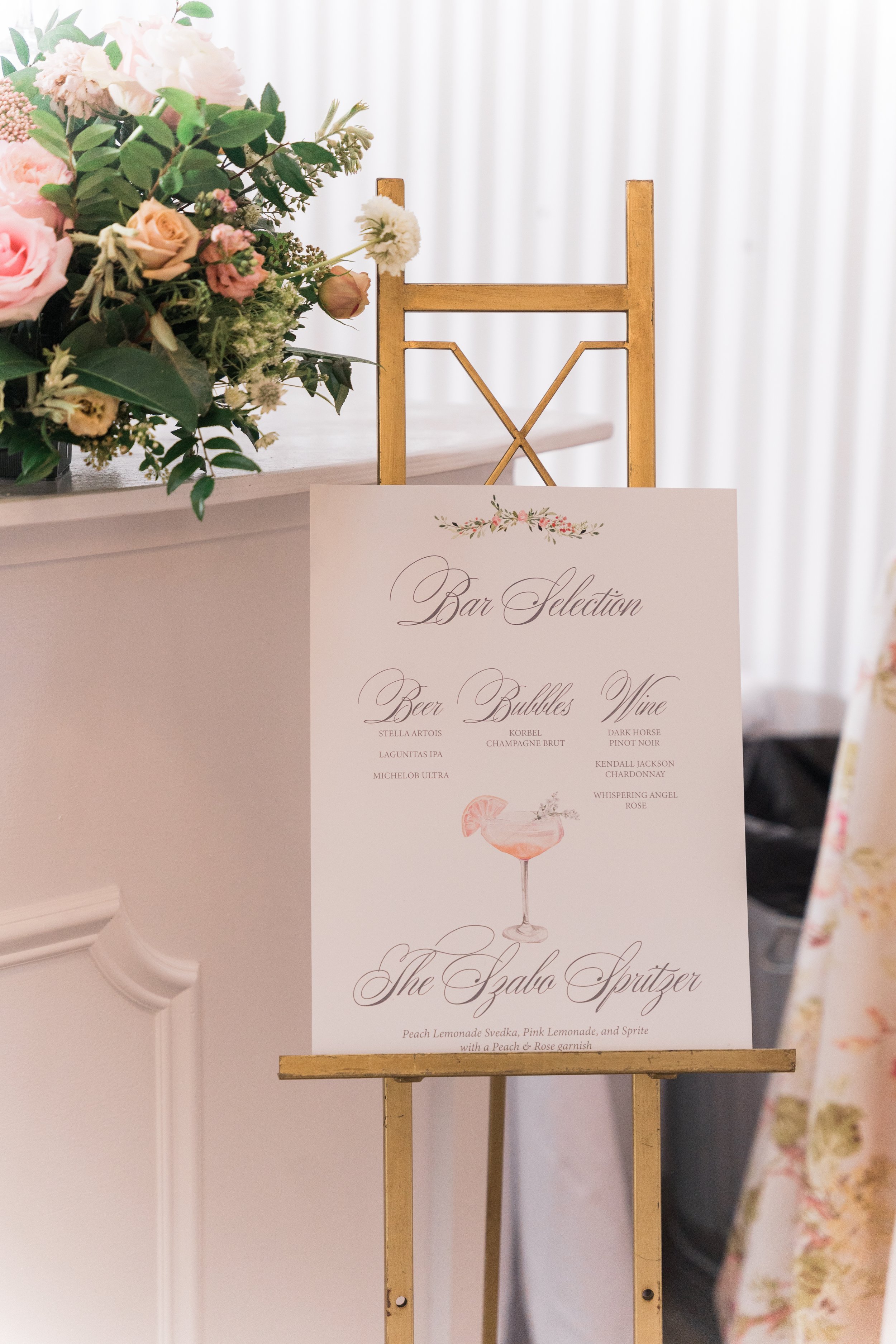 Elegant wedding bar menu on a white poster with floral accents displayed on a gold easel, positioned next to a bouquet of pink and white flowers.