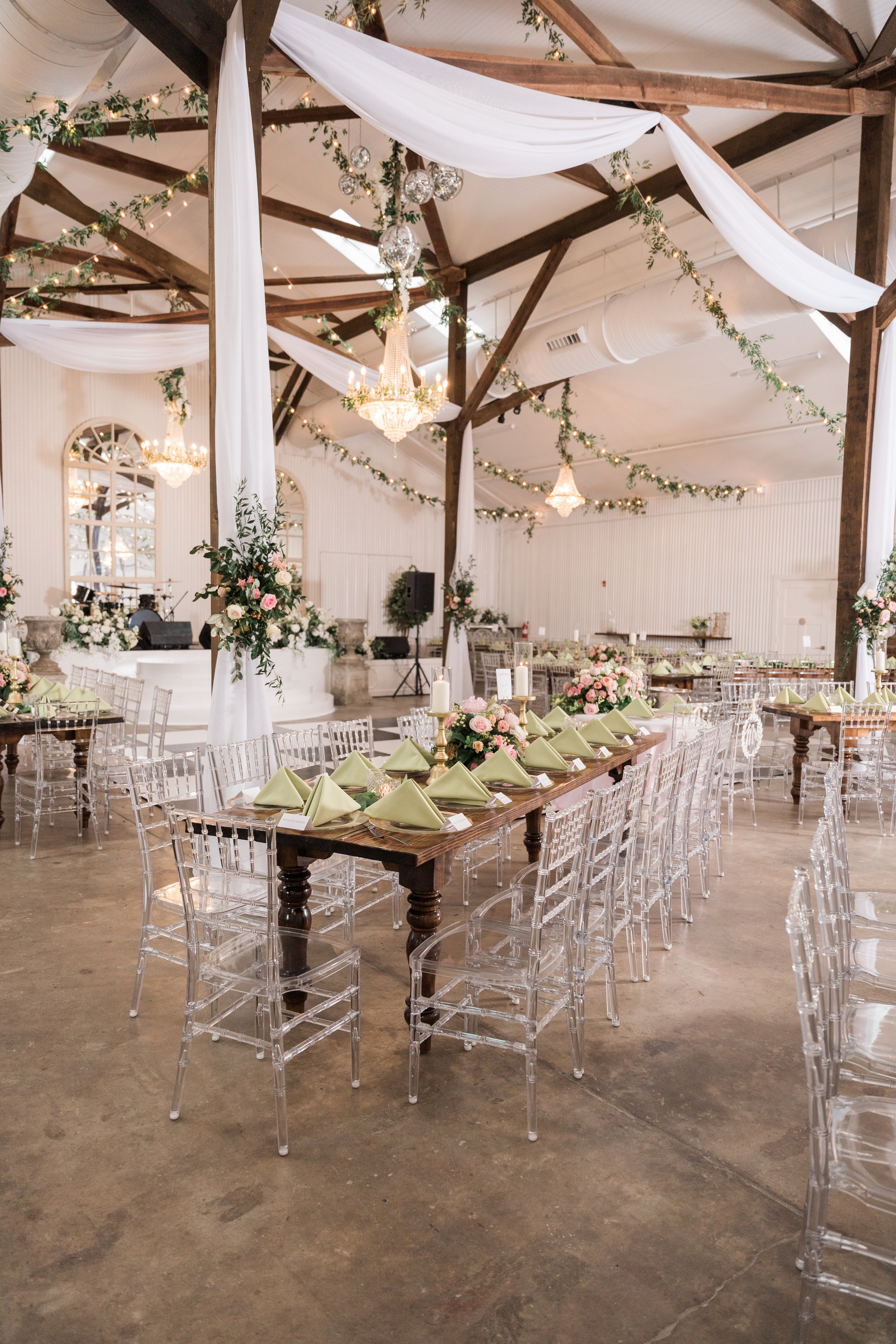 Pensacola Wedding reception with long wooden table set with green napkins, pink and white floral arrangements, candles, and clear chairs, decorated with draped white fabric, chandeliers, and greenery in a rustic venue.