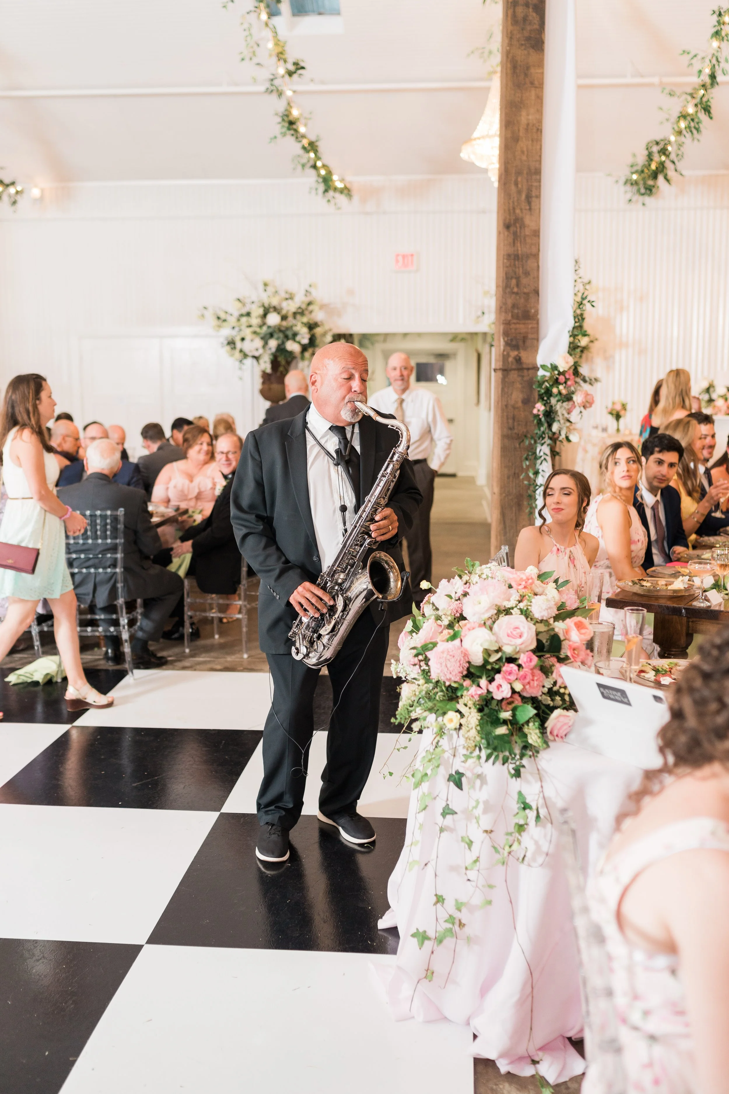 A man in a tuxedo playing a saxophone at a wedding reception with guests seated at decorated tables and floral arrangements.