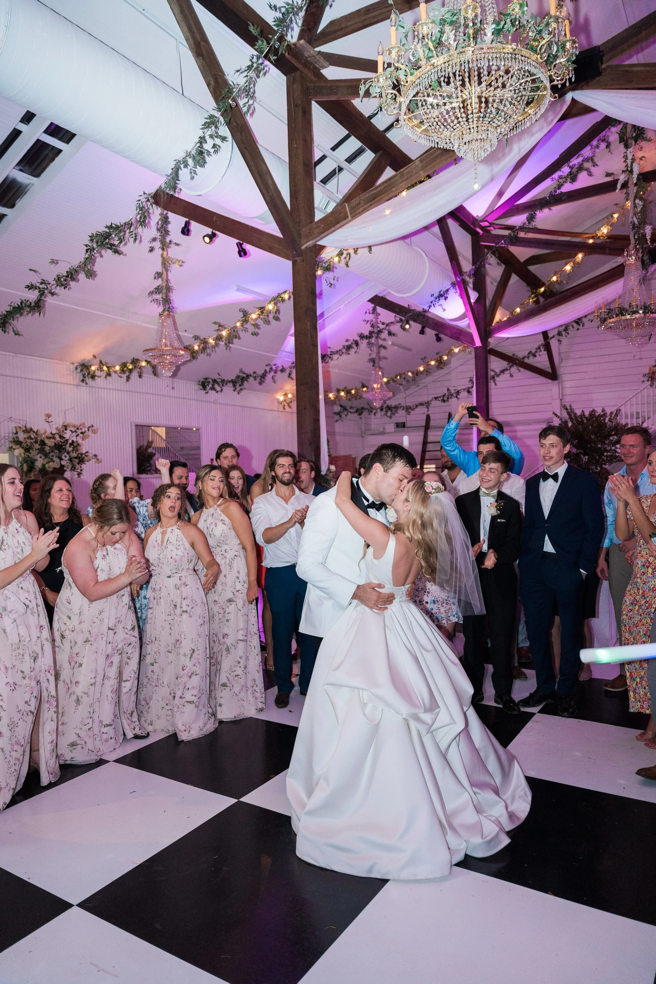 A bride and groom are dancing in the center of a wedding reception, surrounded by guests clapping and smiling. The venue has a rustic wooden ceiling with hanging chandeliers, string lights, and decorative greenery.